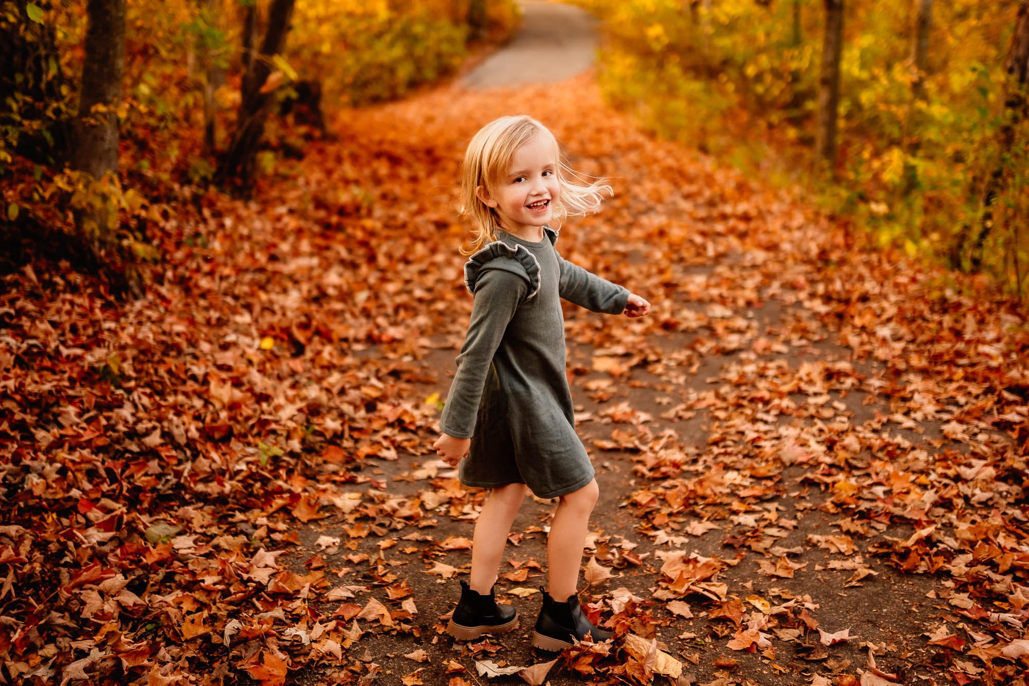 A young girl in a green dress smiling and twirling on a path covered with autumn leaves, surrounded by vibrant golden and orange foliage