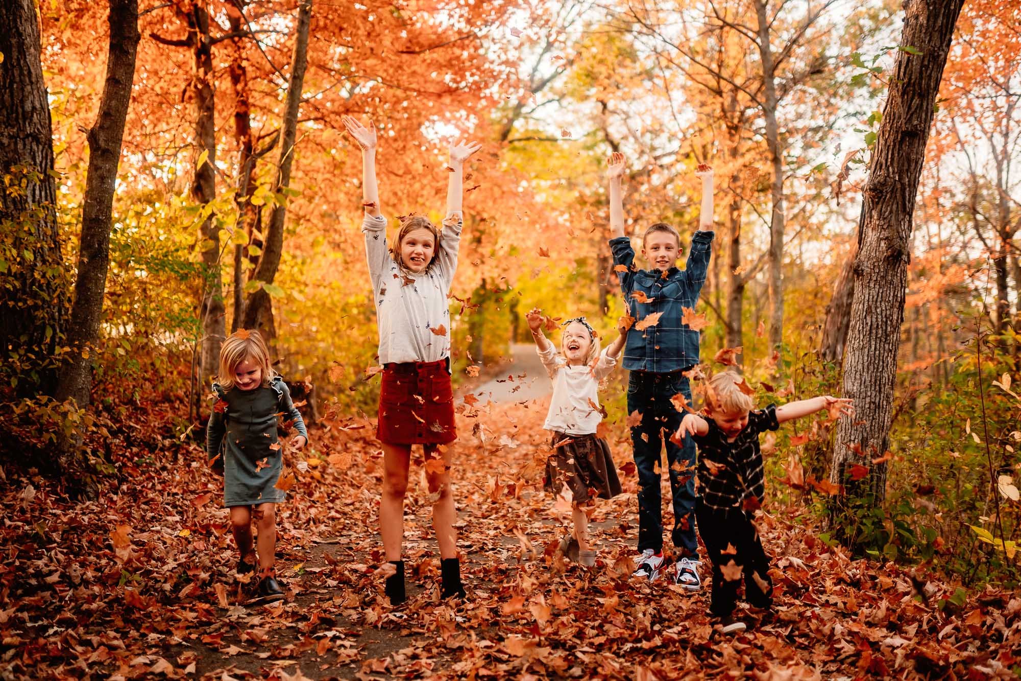Group of children joyfully throwing autumn leaves into the air on a forest path surrounded by vibrant orange and yellow fall foliage