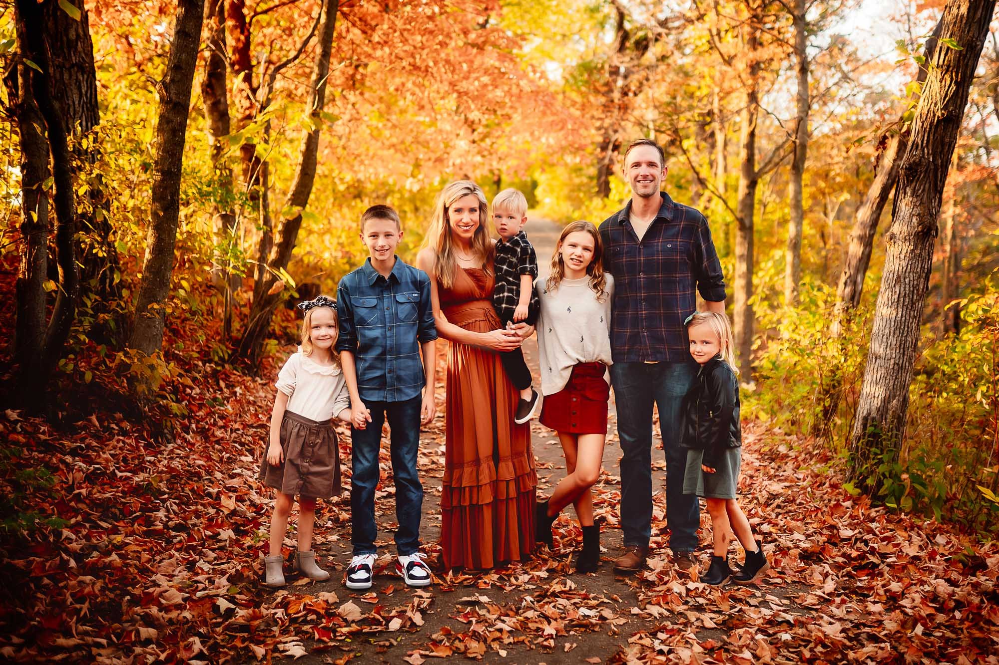 A family of seven standing together on a leaf-covered path surrounded by vibrant fall foliage, smiling and enjoying the warmth of an autumn day
