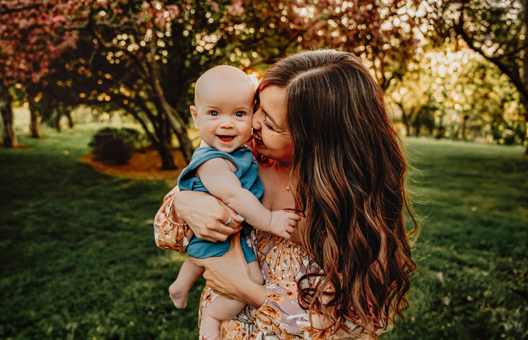 smiling baby held by mom in a floral dress, surrounded by cherry blossoms and golden hour light