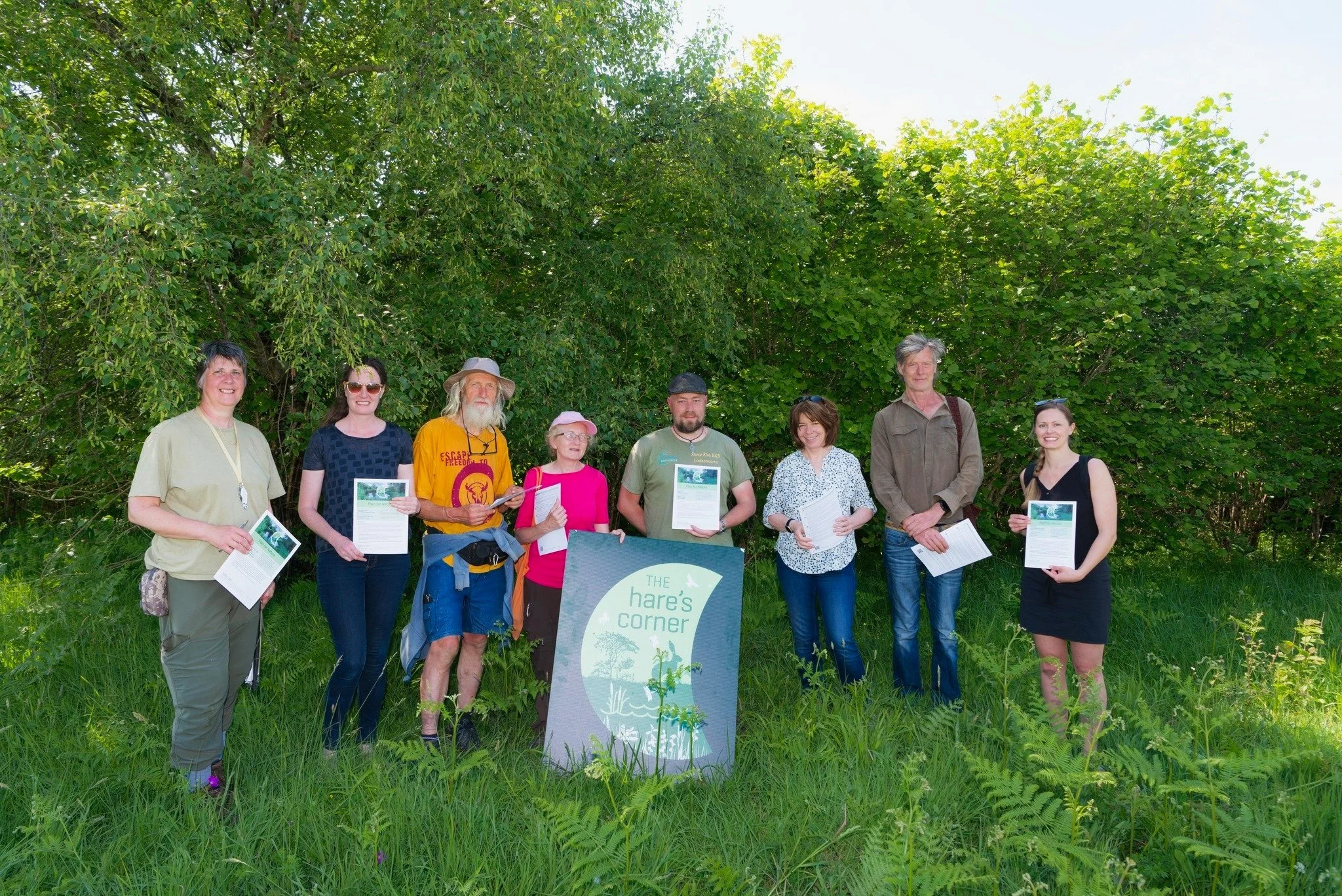 A group of volunteers posing together with a sign that reads “Hair Corners Project.” Some hold informational leaflets, smiling proudly in a natural outdoor setting.
