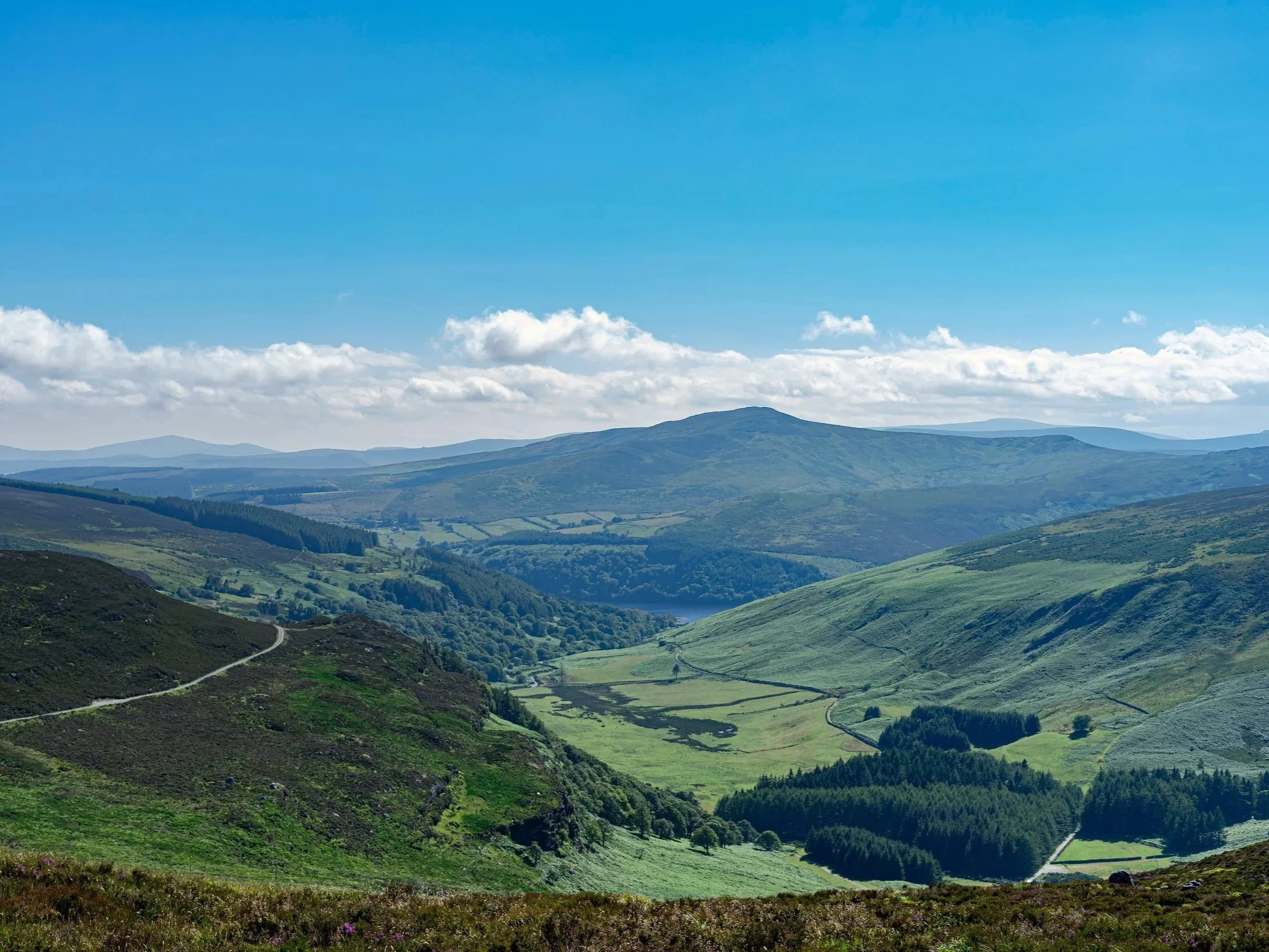 A broad Irish valley with green hills, patchwork fields, and areas of woodland under a blue sky.