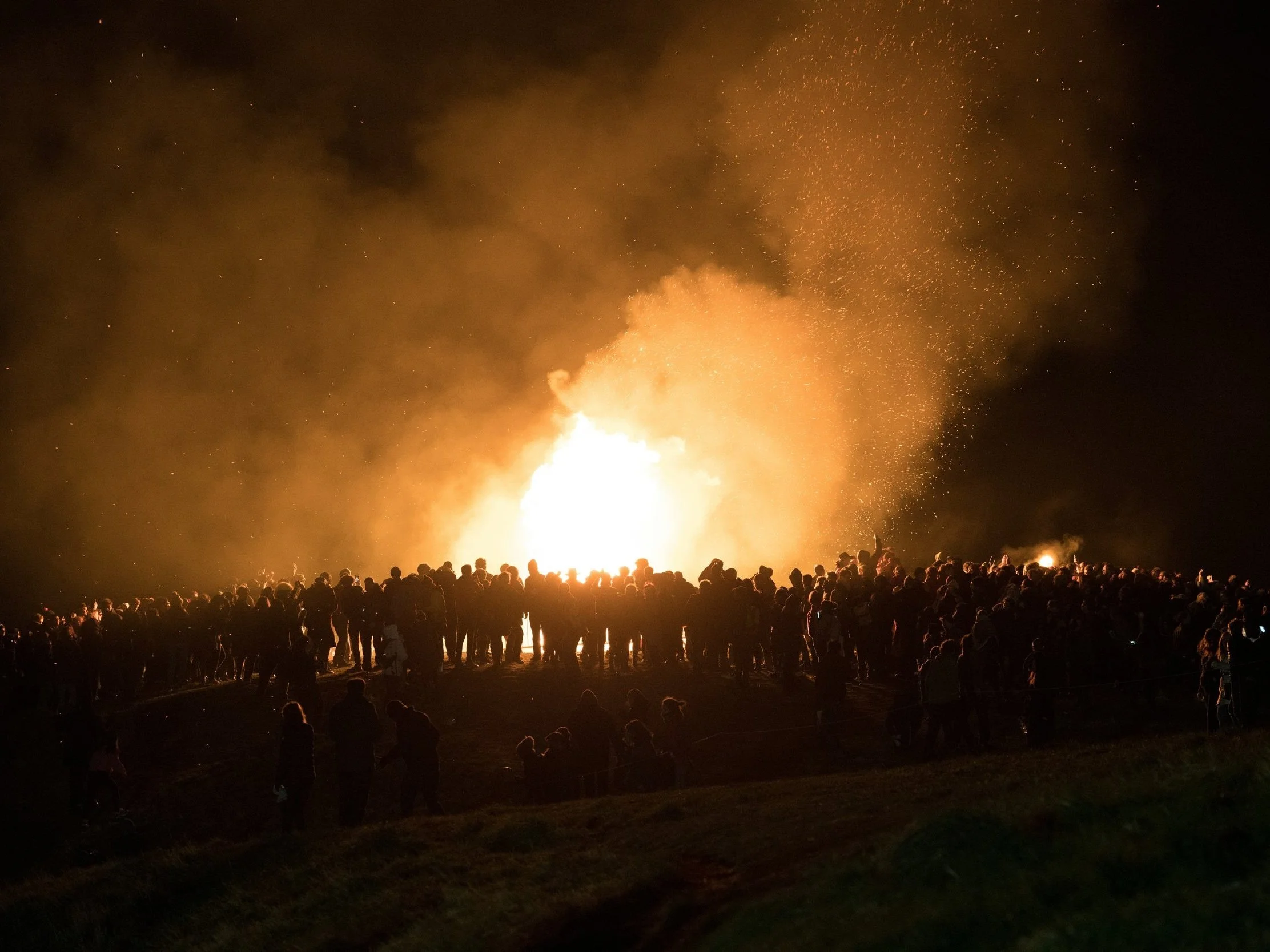 Crowd encircling a large bonfire during an Irish fire festival at night, with sparks rising into the dark sky.