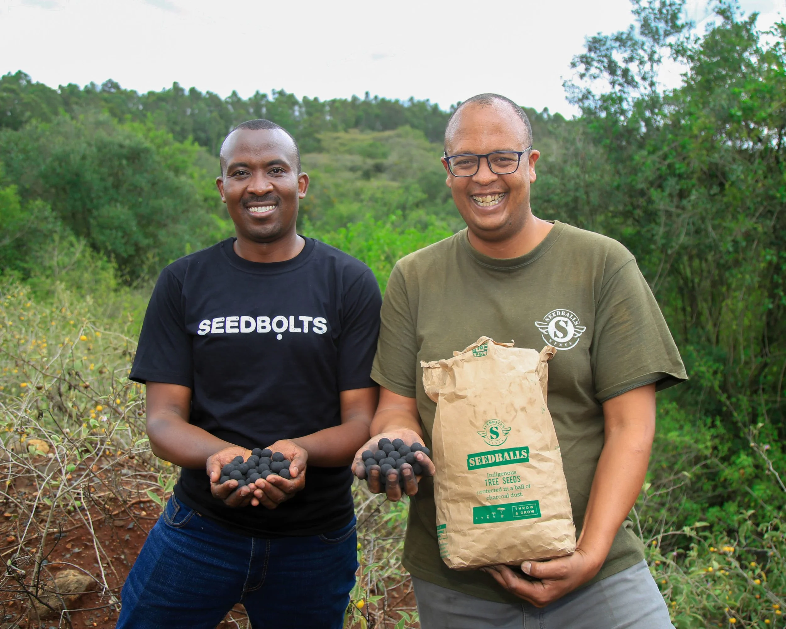 Two smiling men outdoors holding seedballs, one with a bag of seedballs, with green hills and trees in the background.