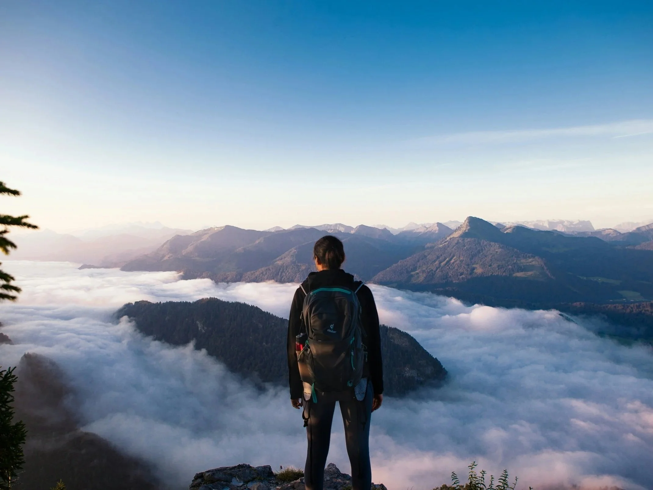 Hiker standing on a mountain summit above clouds at sunrise overlooking ancient mountain landscape.