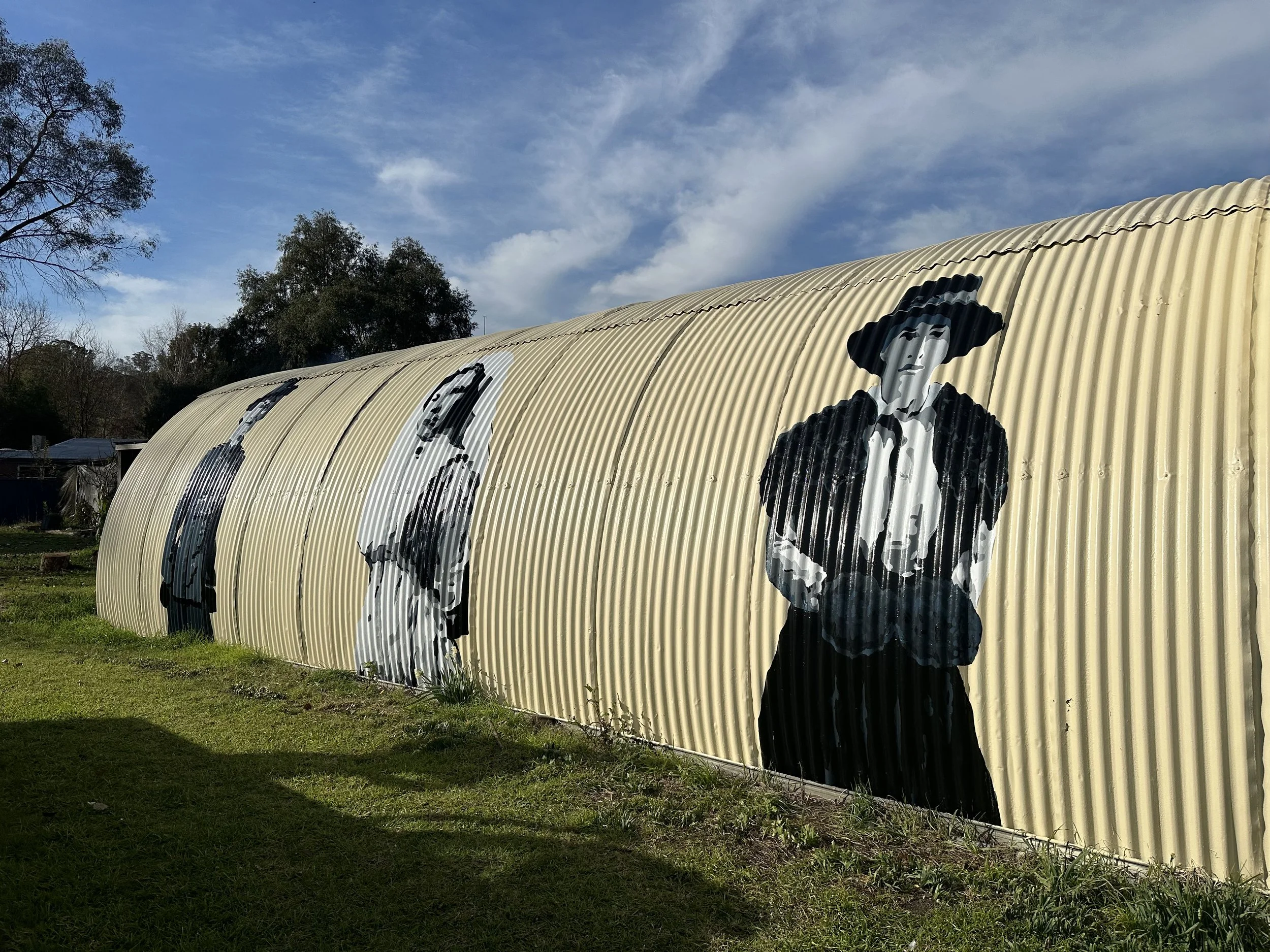 Street art graffiti murals on a corrugated metal wall depicting stylized figures, including a woman with a large hat, against a clear blue sky with some clouds, and green grass in the foreground.