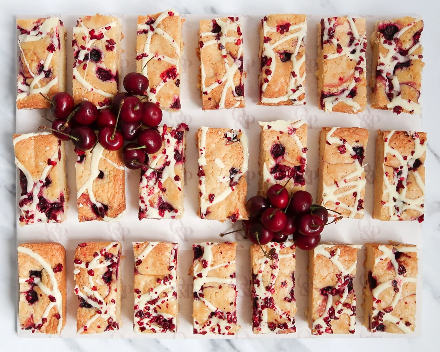 Morello cherry blondies topped with white chocolate drizzle, garnished with fresh cherries, arranged on a white platter. Blondies.