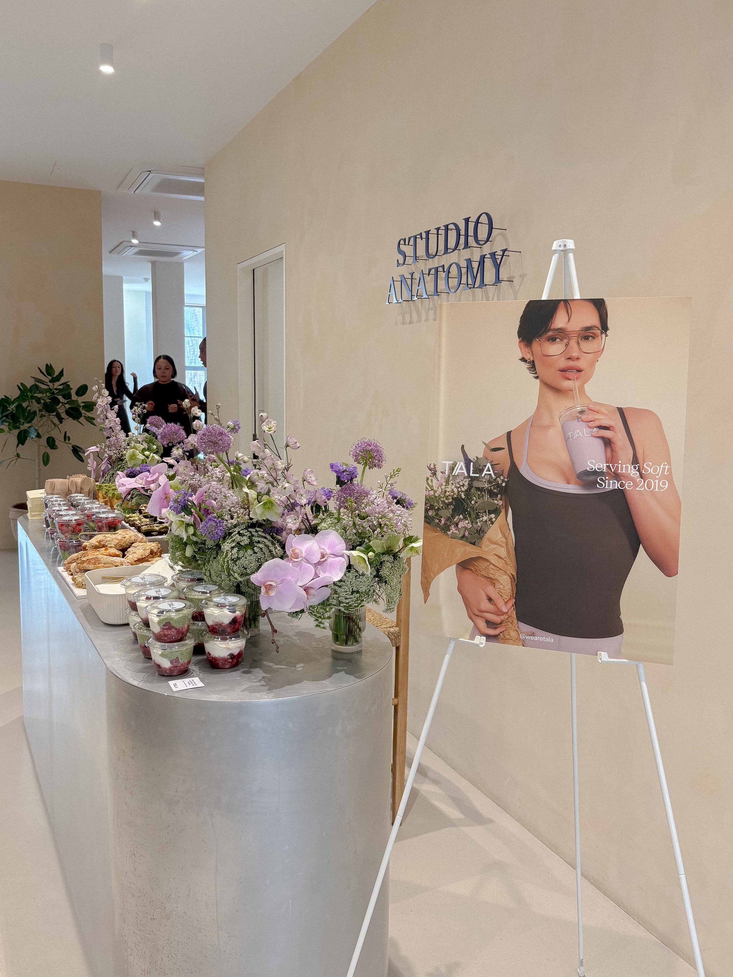 A reception table with various desserts and a floral arrangement, next to a large poster of a woman holding a flower bouquet and drinking from a cup with a straw, inside a modern, well-lit room with a sign reading 'Studio Anatomy' on the wall.