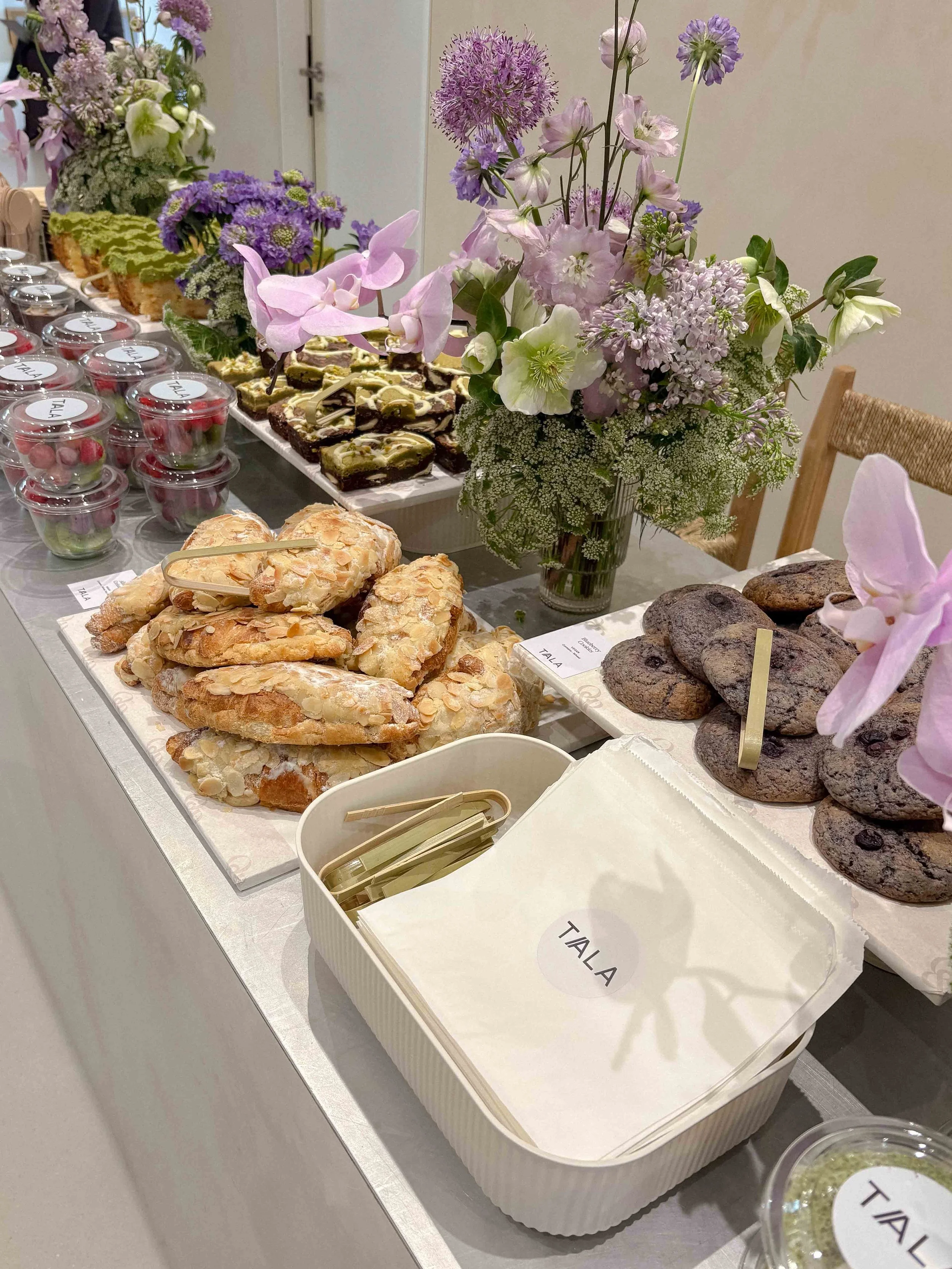 A table with an assortment of baked goods including cookies, pastries, and small desserts, decorated with a large bouquet of pastel purple and pink flowers, and labeled with a sign that says 'TALA'.