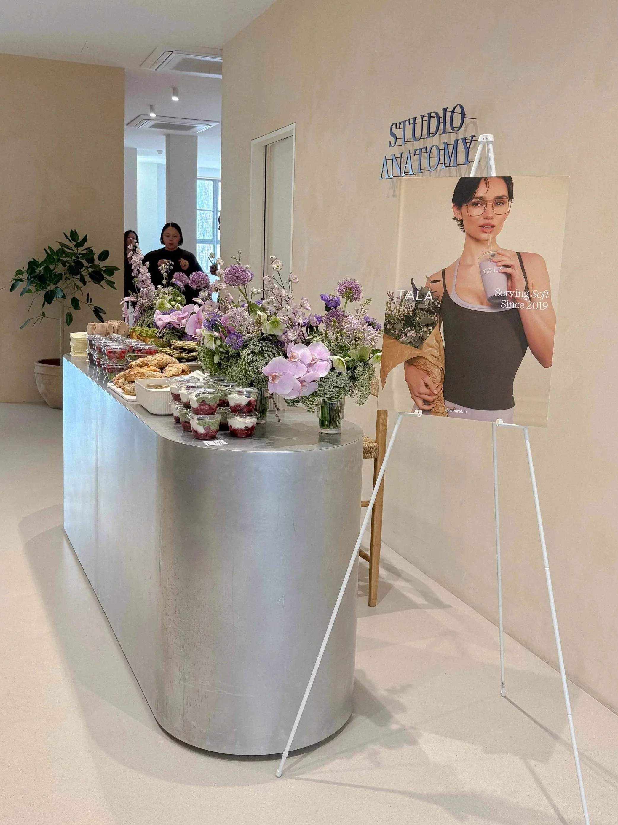 Display table with pink and purple flowers, assorted pastries, and breakfast pots, with a promotional poster of a woman holding a drink in the background at Studio Anatomy.