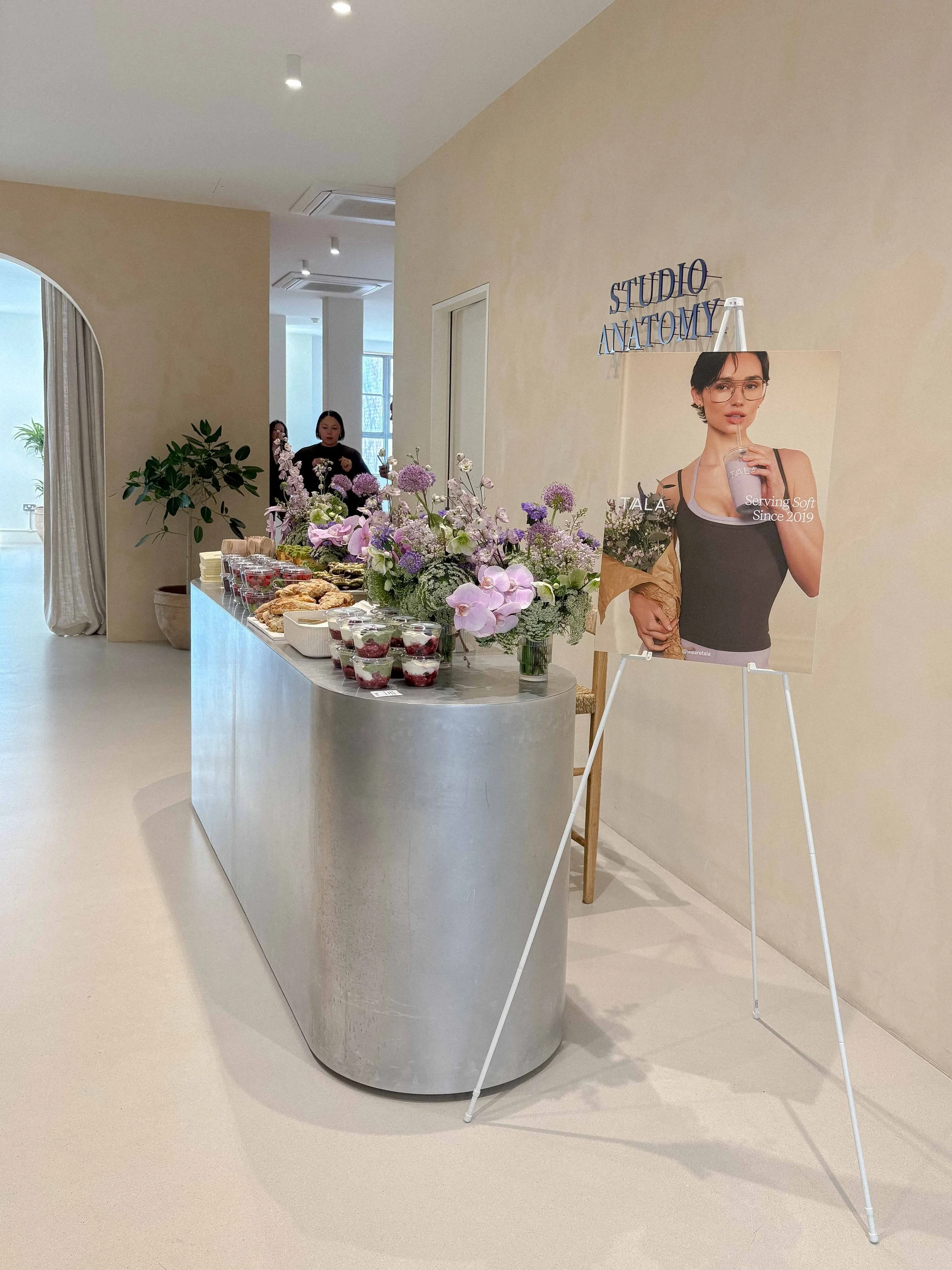 A reception desk with pink and purple flowers and assorted desserts and snacks, a portrait of a woman holding a drink and wearing glasses, and two women standing in the background in front of a window, inside a room labeled 'Studio Anatomy'.