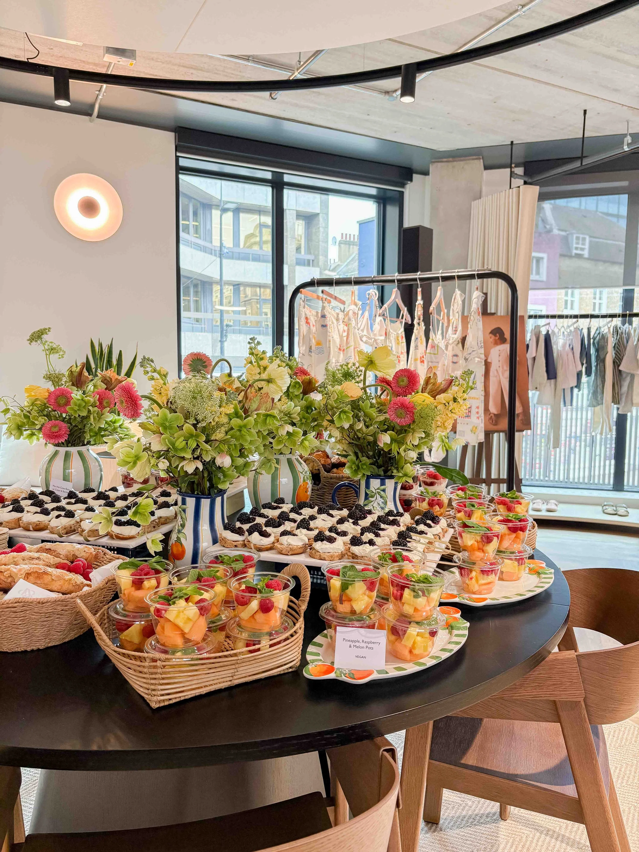 Display table with pink and purple flowers, assorted pastries, and breakfast pots, with a promotional poster of a woman holding a drink in the background at Studio Anatomy.