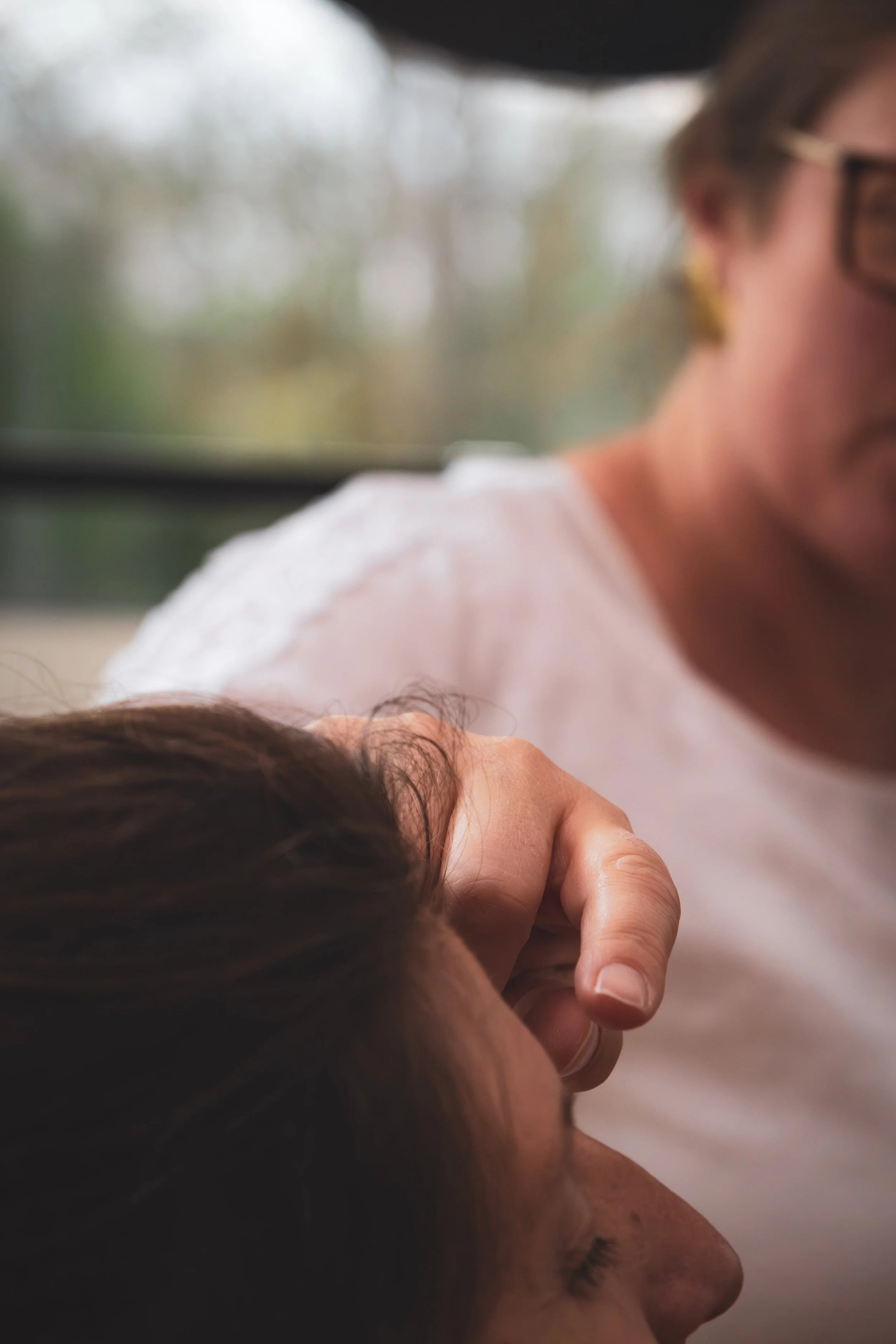 Une personne pose sa main sur le visage d'une autre personne allongée, en intérieur, avec un arrière-plan flou d'un paysage naturel.