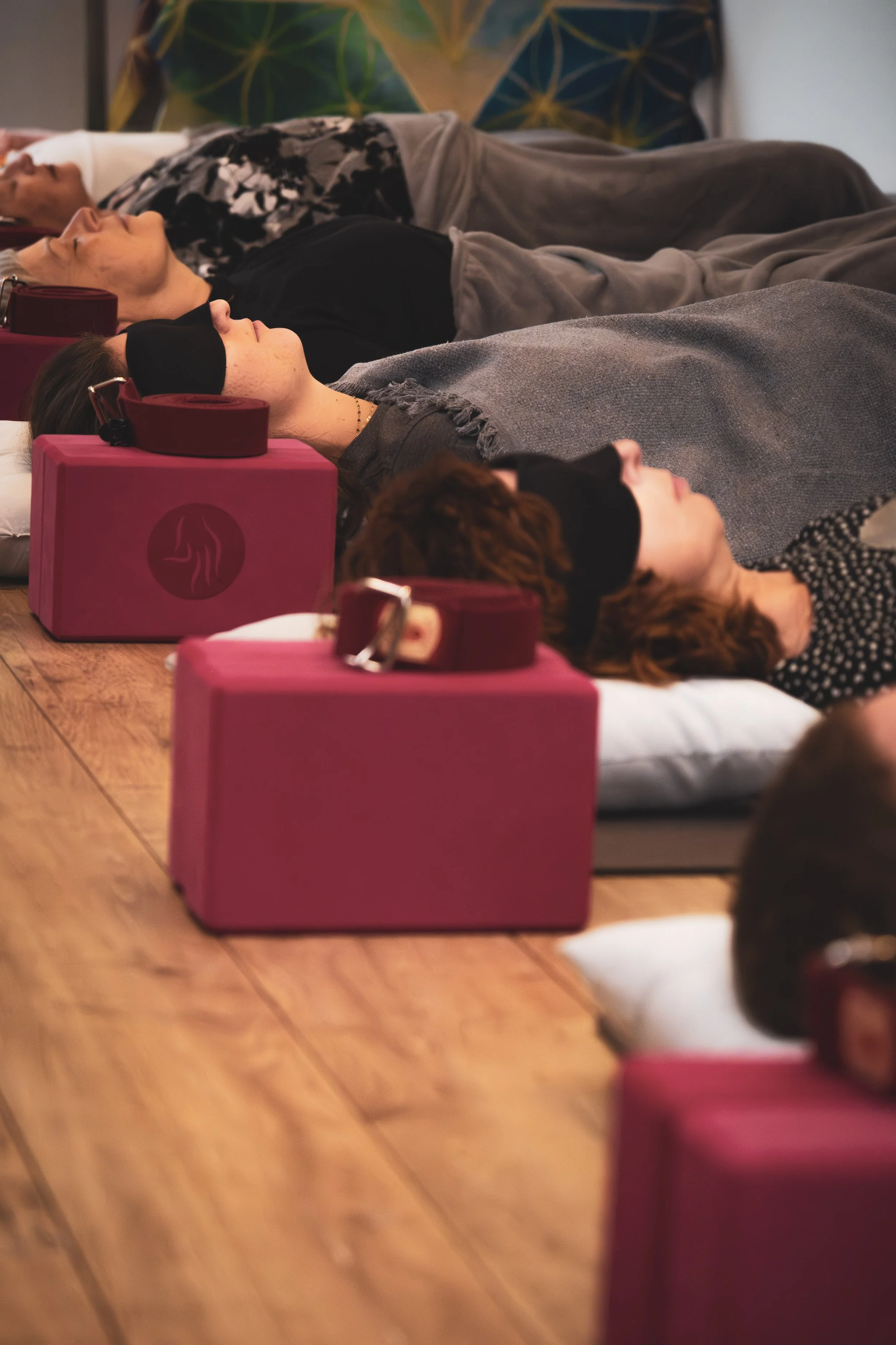 Groupe de personnes allongées sur le sol, portant des masques pour dormir ou méditer, avec des blocs de yoga rose et des bandes de résistance à côté, dans une salle de spa ou de yoga.
