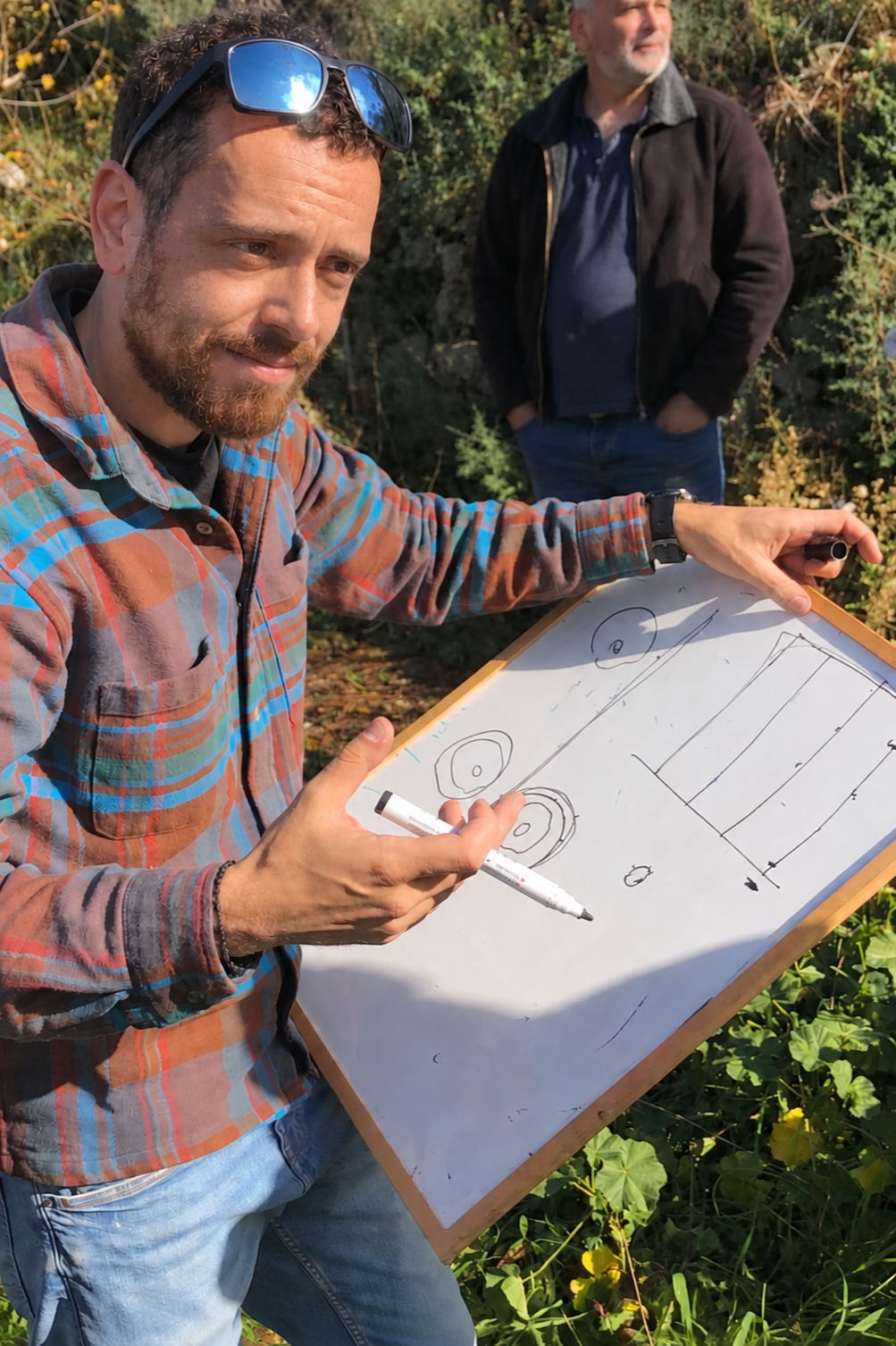 A man with sunglasses on his head making drawings on a whiteboard, with another man standing behind him outdoors among greenery.