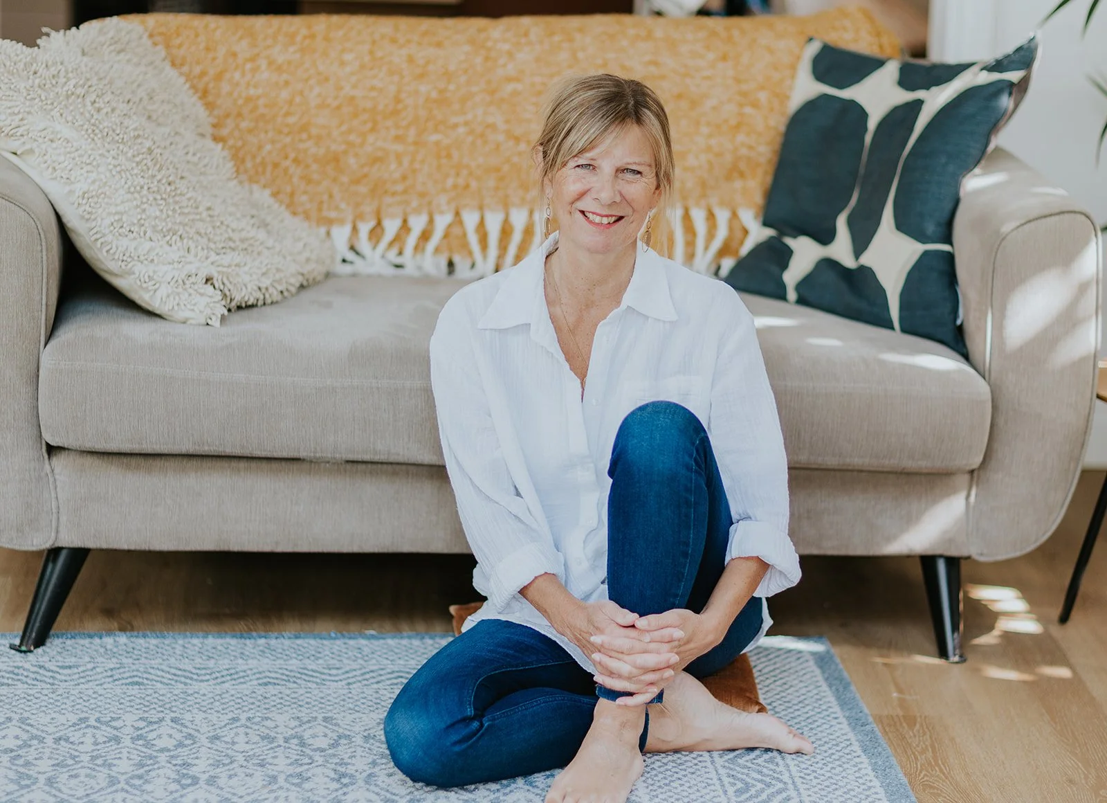 A woman sitting on the floor in front of a beige sofa, smiling, with one knee up and her hands clasped around her leg. The sofa has a textured cream pillow, an orange throw blanket, and a dark blue pillow with white abstract patterns. The room has na