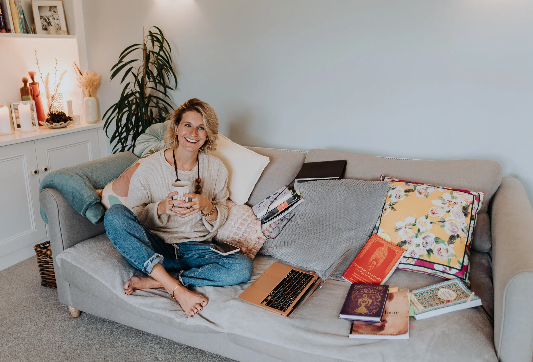 A woman sitting on a beige couch with books, a laptop, and a phone, holding a mug and smiling in a cozy living room.
