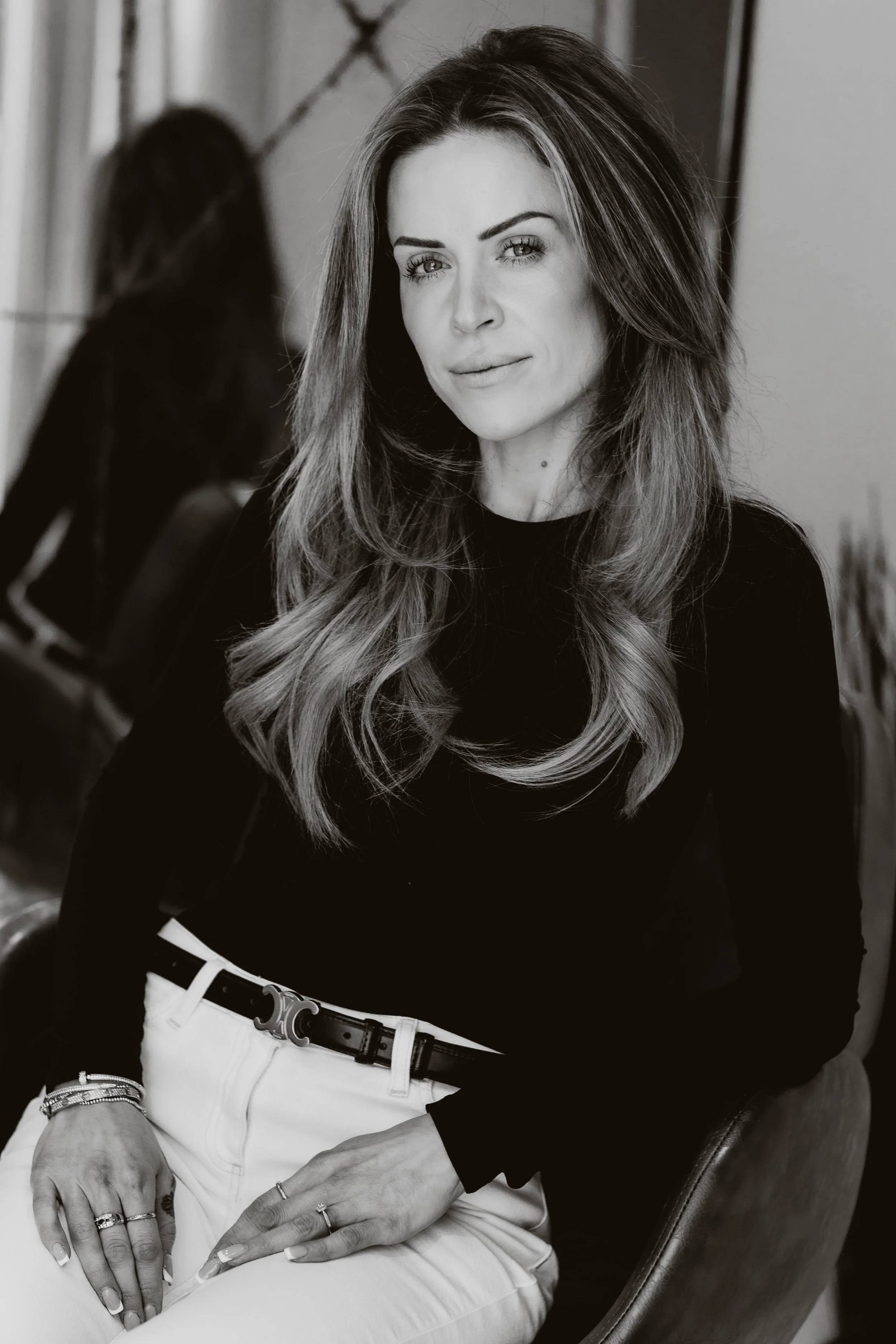 A woman with long, wavy hair sitting in a chair, wearing a black top and white pants, looking at the camera in a professional setting.