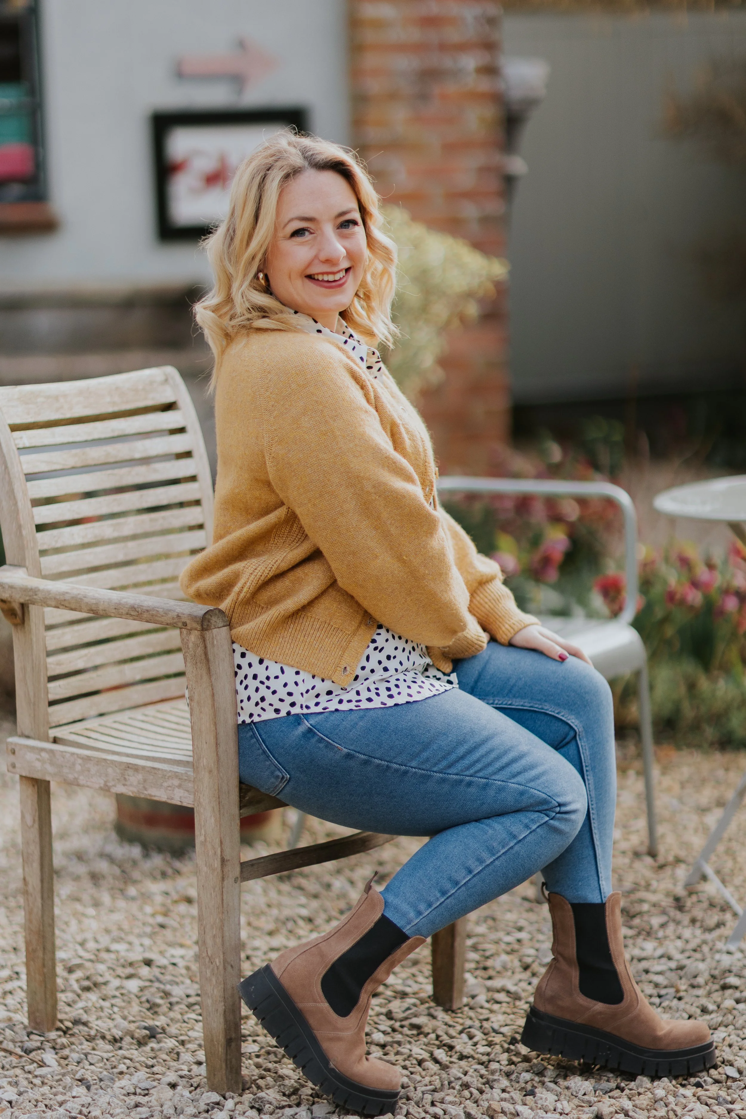 A woman with blonde hair and a warm smile sitting on a wooden bench outdoors, wearing a tan cardigan, a white polka-dot blouse, blue jeans, and brown boots, with a garden and brick wall in the background.