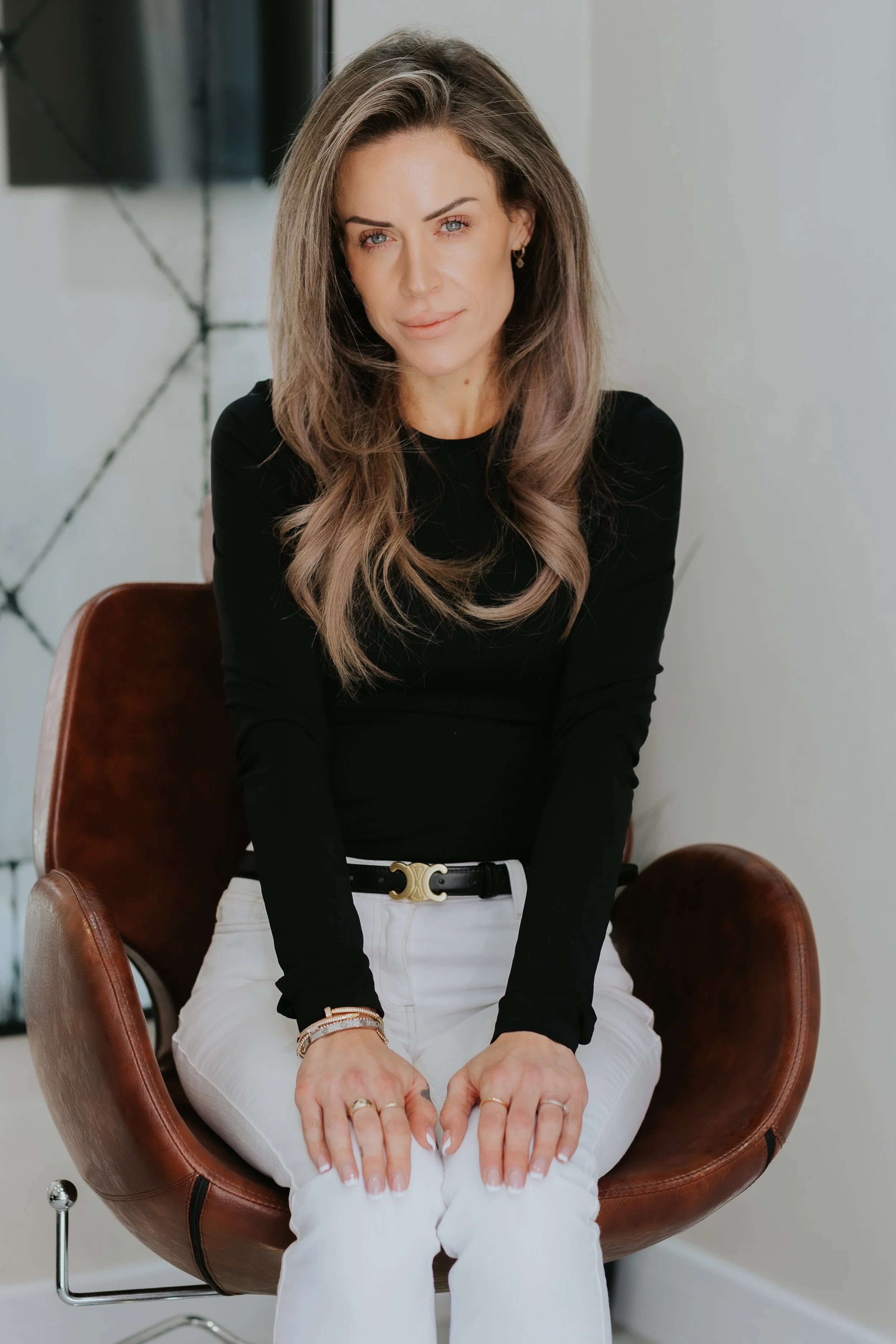 A woman with long, light brown hair sits on a brown leather chair in a modern room, wearing a black long-sleeve top and white pants.