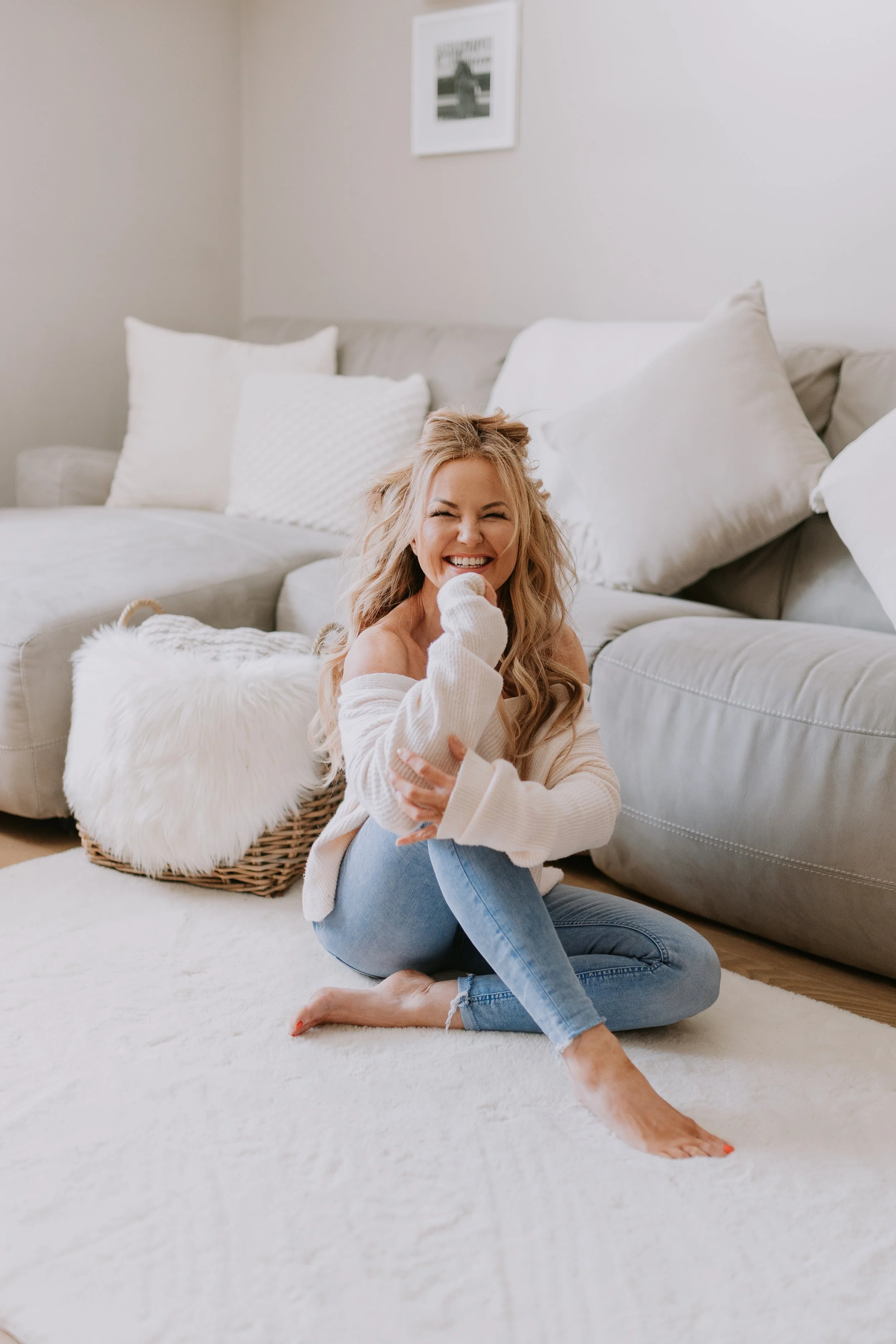 A woman with long blonde hair, wearing an off-shoulder white sweater and light blue jeans, sitting on a cream-colored rug in front of a gray couch, smiling and laughing.