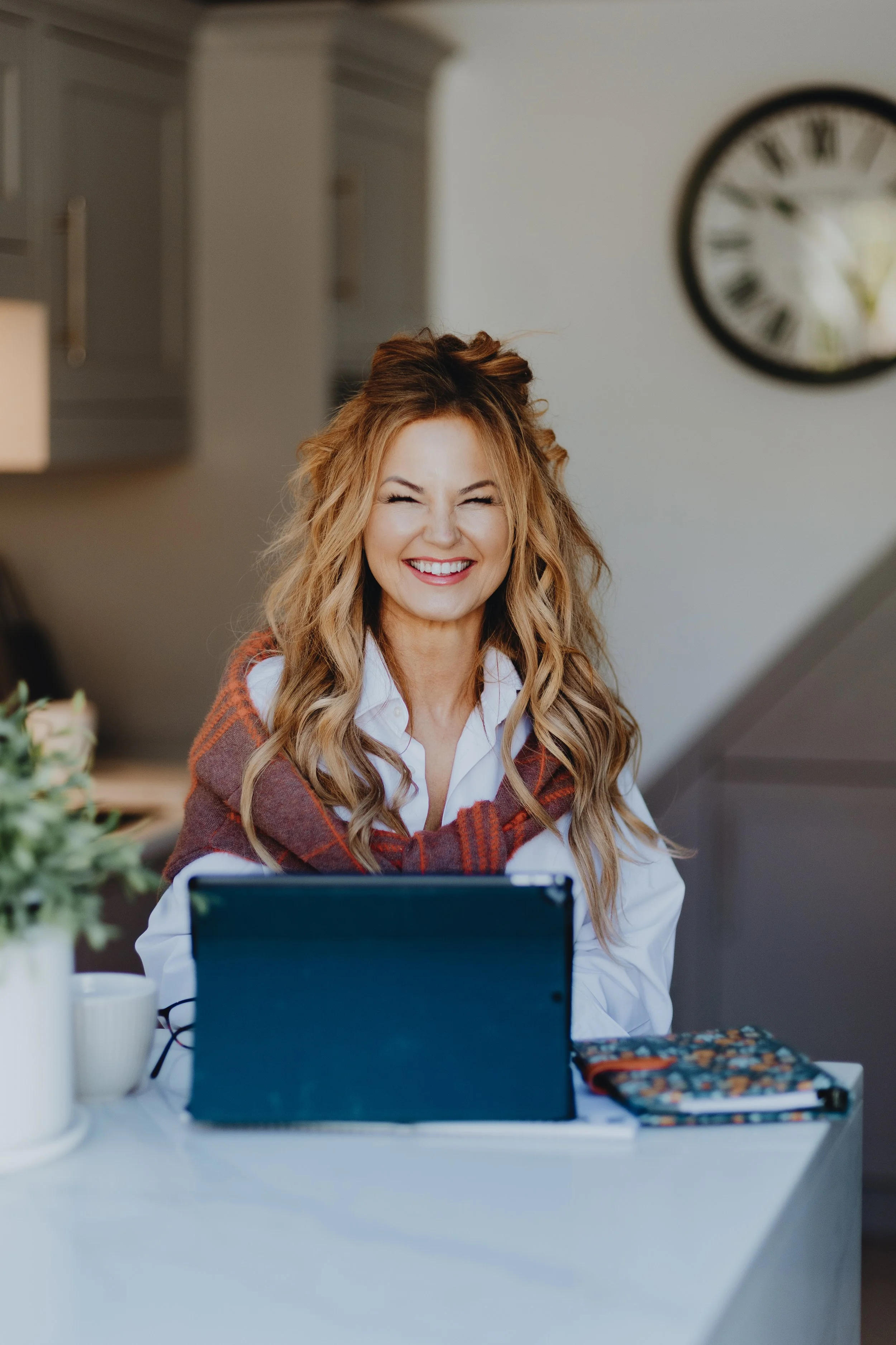 Smiling woman with long wavy hair sitting at a kitchen table with a laptop, notebook, and mug, in a cozy modern kitchen.