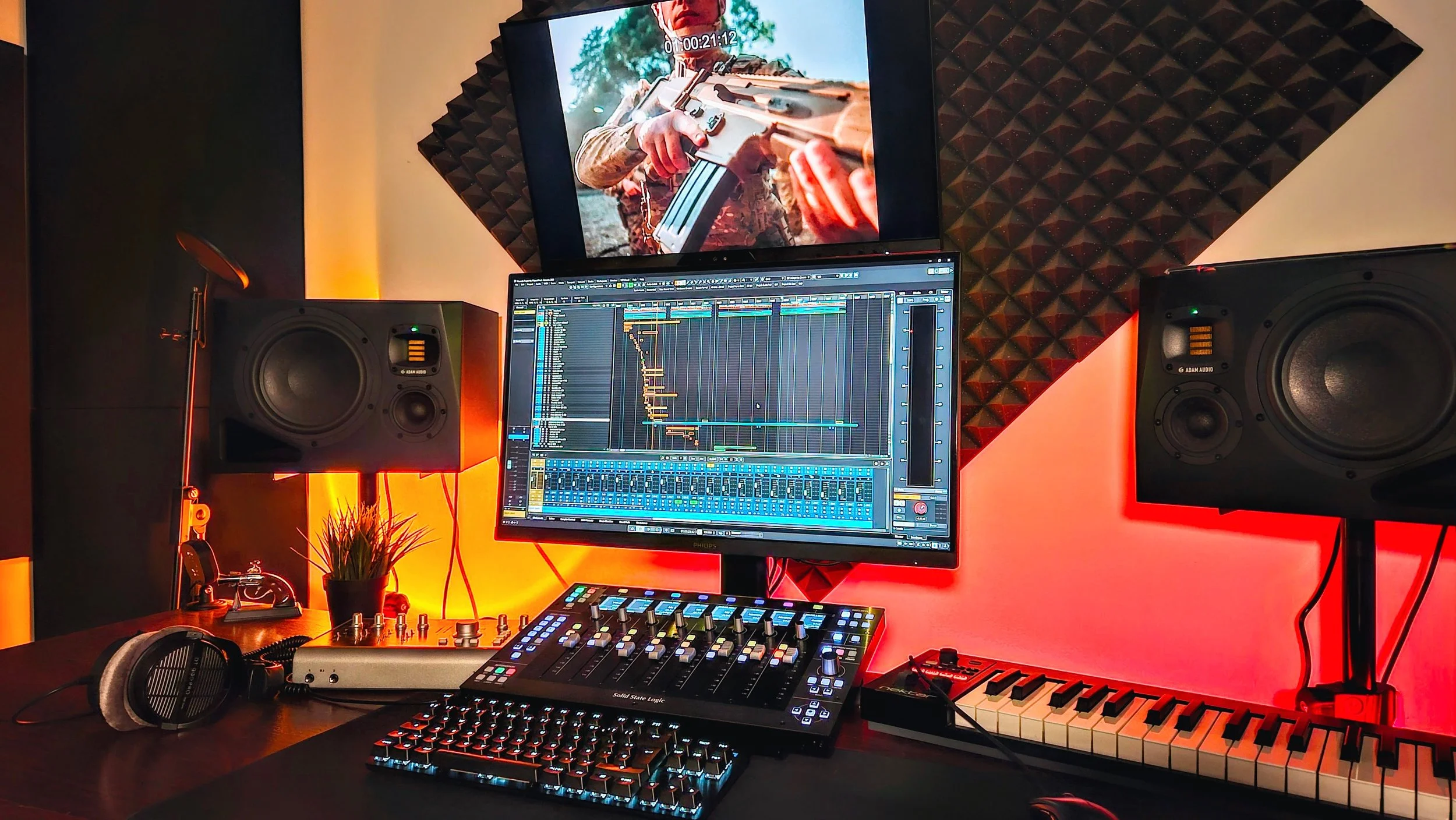 A music production and recording studio with a computer displaying music editing software, studio monitors, a MIDI keyboard, a mixer, headphones, and a large screen showing a person holding a rifle.