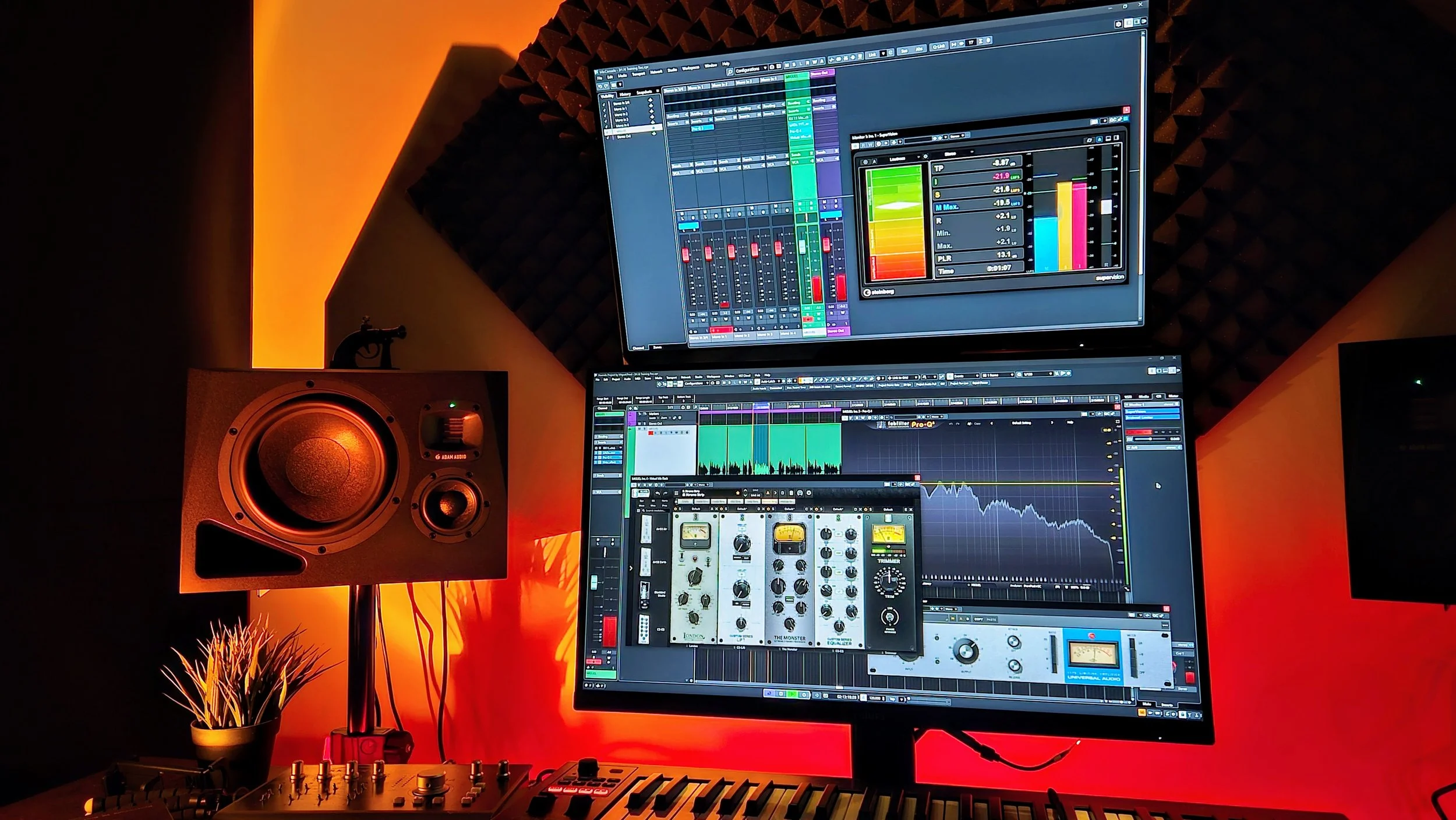 A music production studio with dual computer monitors displaying audio editing software, a studio speaker, a small plant, and music equipment on a desk with red ambient lighting.