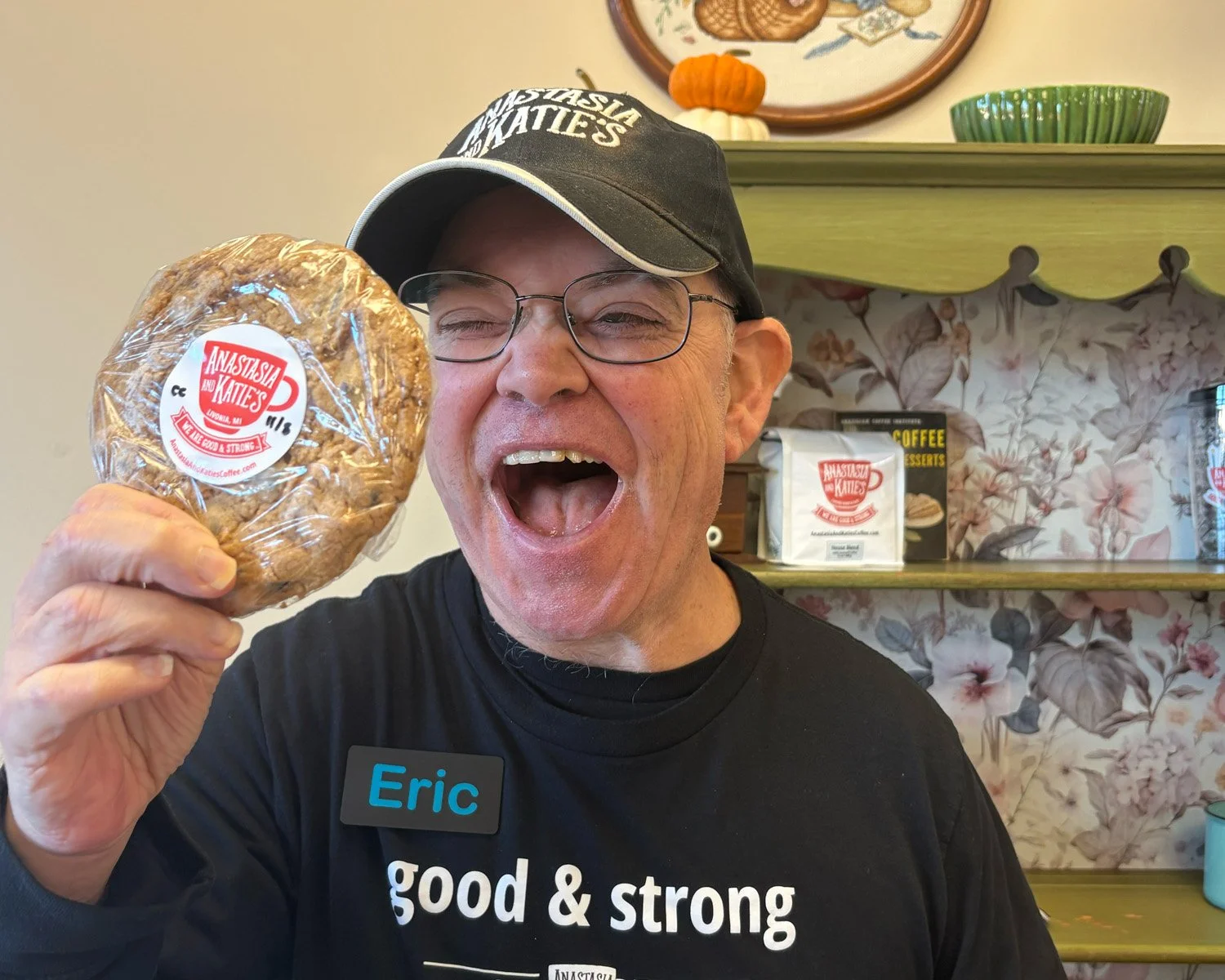 Man with huge, excited smile, holding up a big, homemade cookie with Anastasia and Katie's Coffee Shop sticker logo on it.