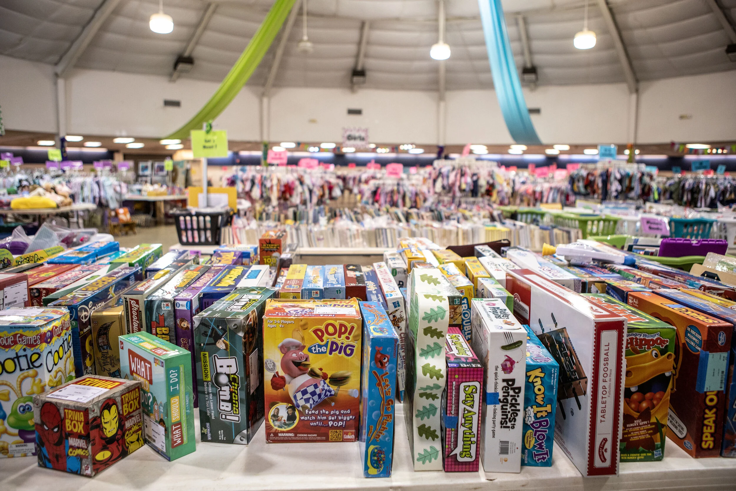 A wide view of a thrift store with shelves filled with board games and children's toys. The store has a high, vaulted ceiling with large hanging lights and colorful streamers.