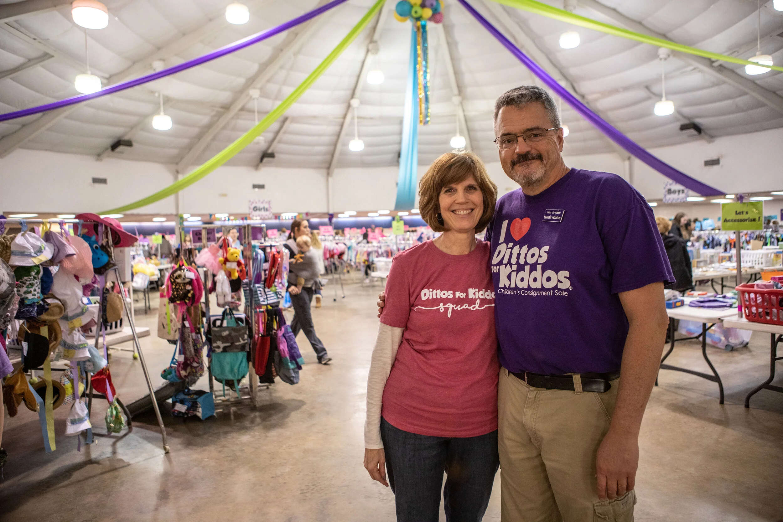 Two people standing in a children's resale store, smiling at the camera. The woman is wearing a pink Dittos for Kiddos squad t-shirt, and the man is wearing a purple Dittos for Kiddos t-shirt with a children’s consignment sale logo. The store is filled with racks of children's clothes, toys, and accessories, with colorful streamers hanging from the ceiling.