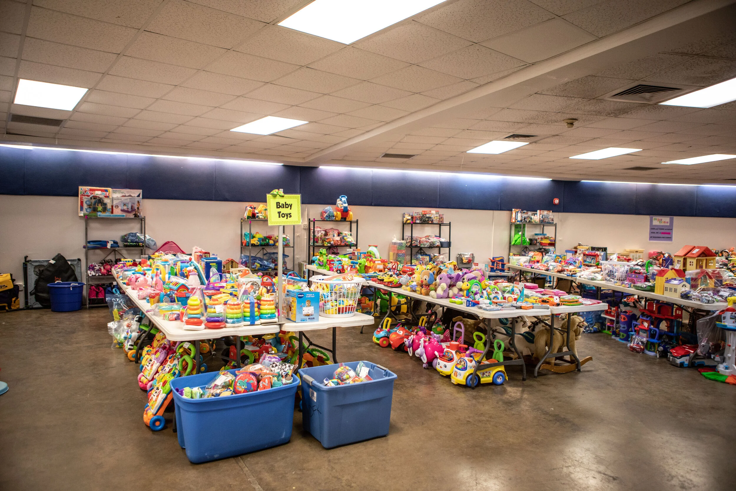 Tables and shelves filled with a variety of children's toys, such as stuffed animals, plastic vehicles, and stacking rings, in a large indoor space with a ceiling of rectangular lights.