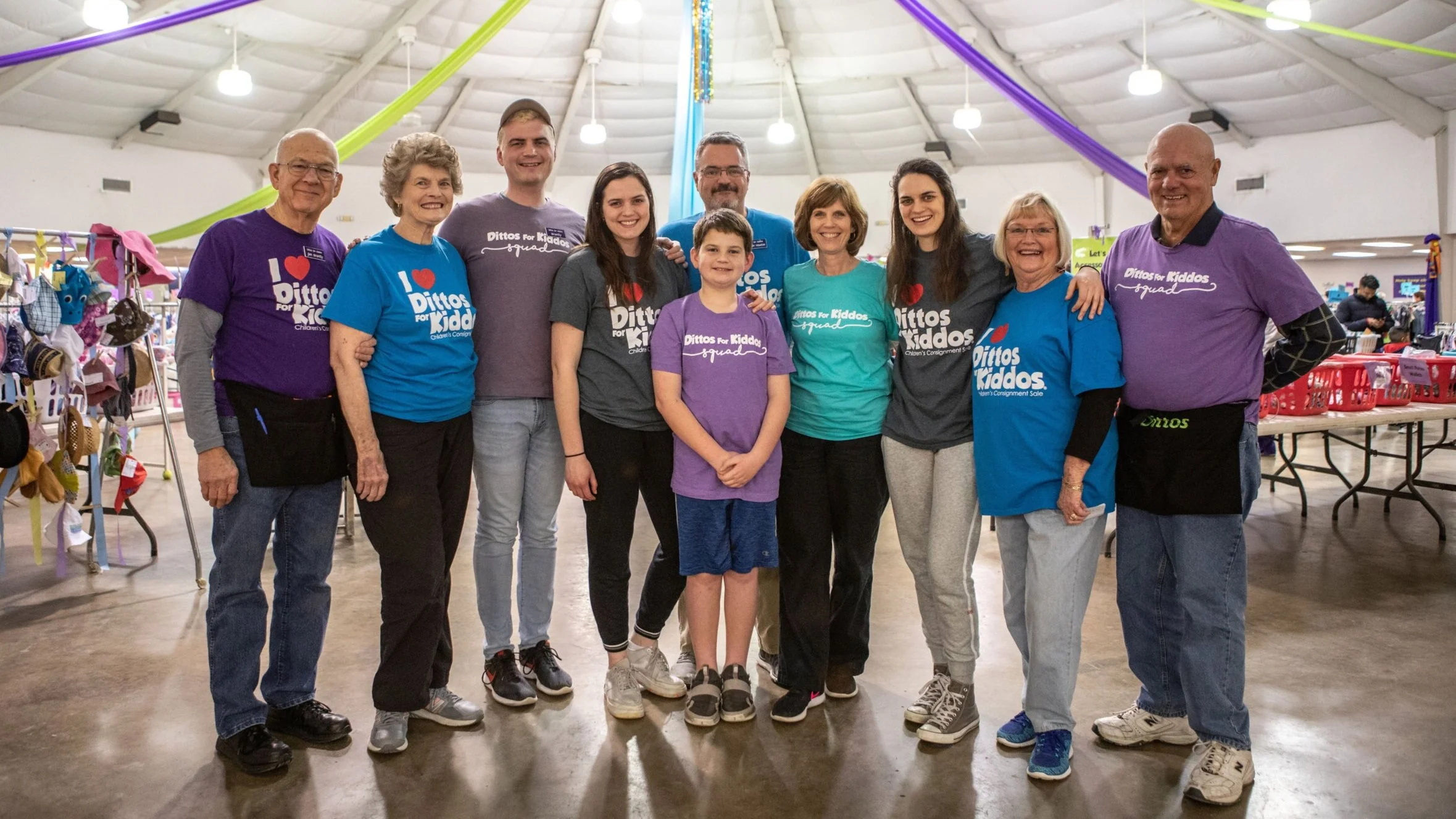 Group of people smiling and standing indoors at a community event, with colorful streamers hanging from the ceiling, and tables with merchandise in the background.