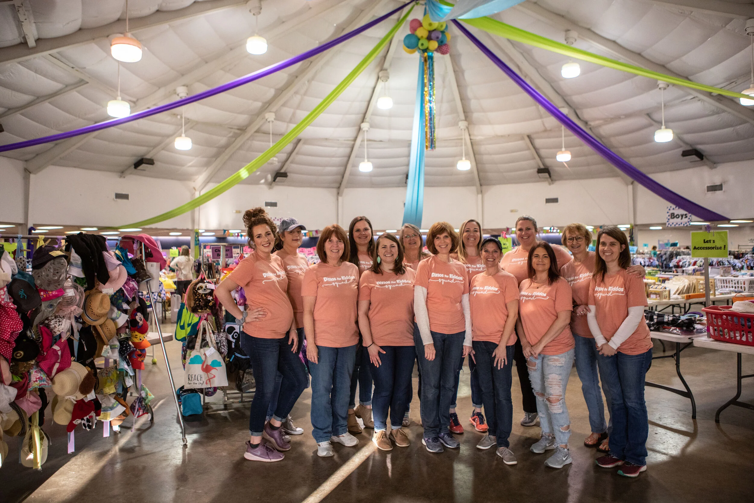 A group of women posing together inside a store decorated with colorful streamers hanging from the ceiling. The women are wearing matching peach T-shirts with the text 'Ditos for Kiddos' and are smiling at the camera. The store's shelves behind them are filled with various clothing, accessories, and hats.