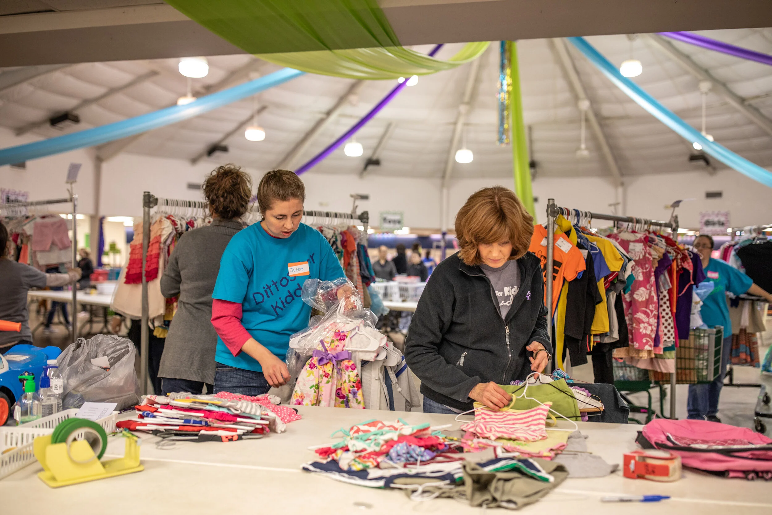 People shopping at a thrift store or donation event, browsing through clothing and accessories on tables and racks under a large indoor space with colorful ceiling decorations.