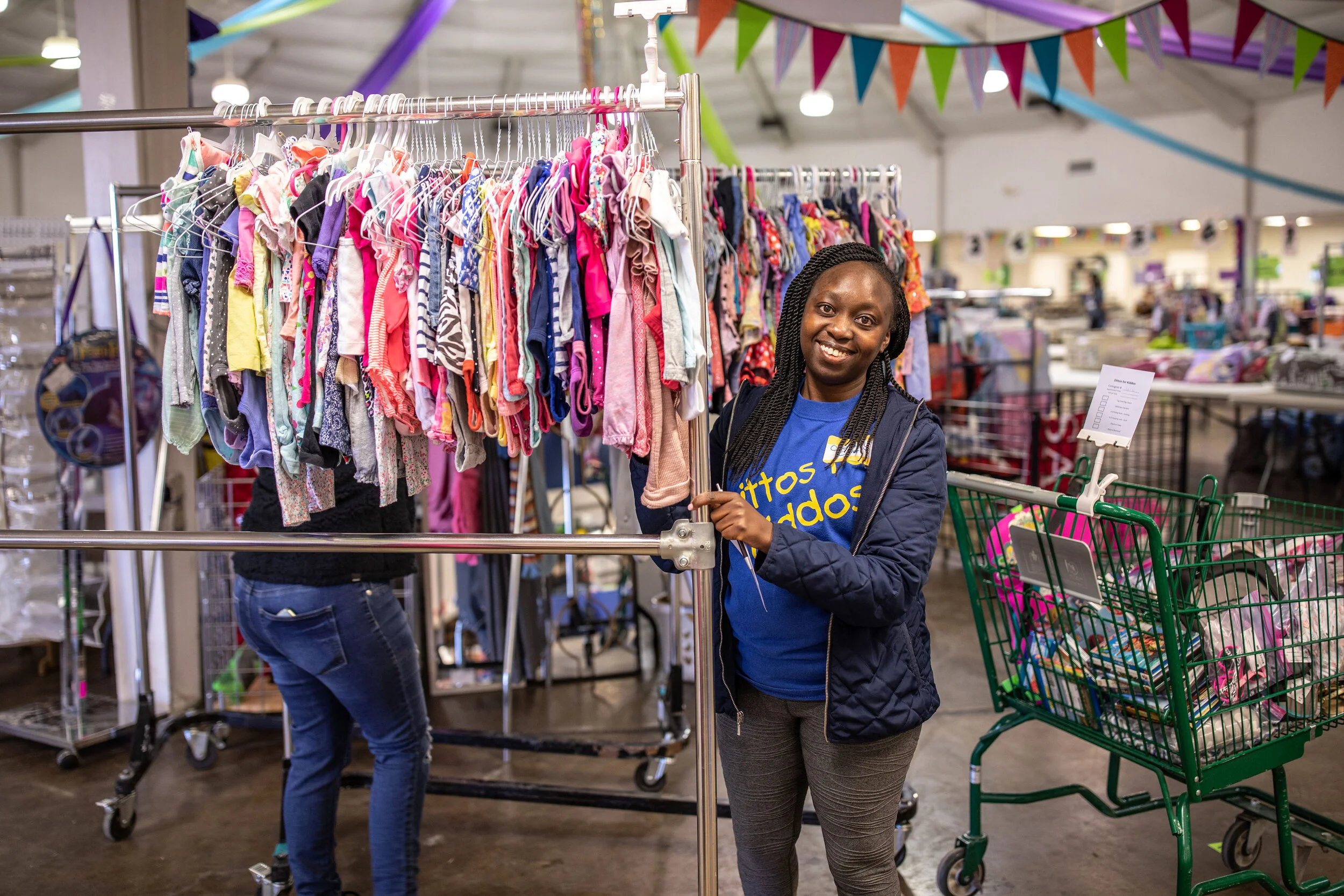 A woman shopping at a thrift store or flea market, standing next to a clothing rack with children's clothes and smiling at the camera. Colorful triangular banners hang overhead in the store.