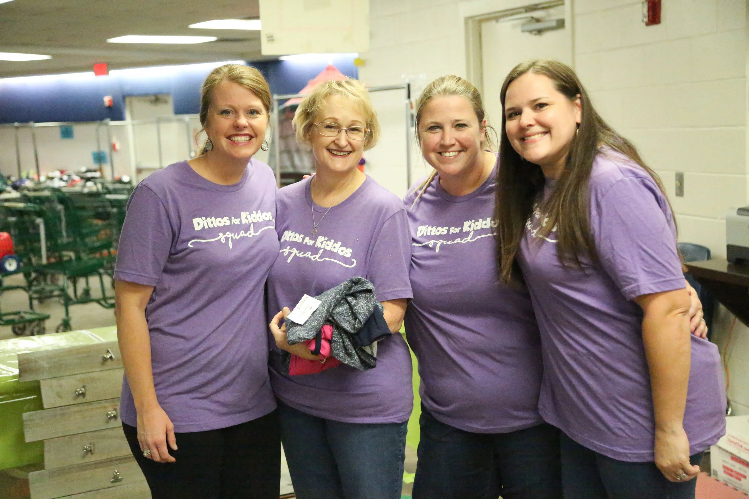 Four women wearing purple T-shirts with 'Ditto for Kiddos Squad' printed on them, smiling and standing together indoors.