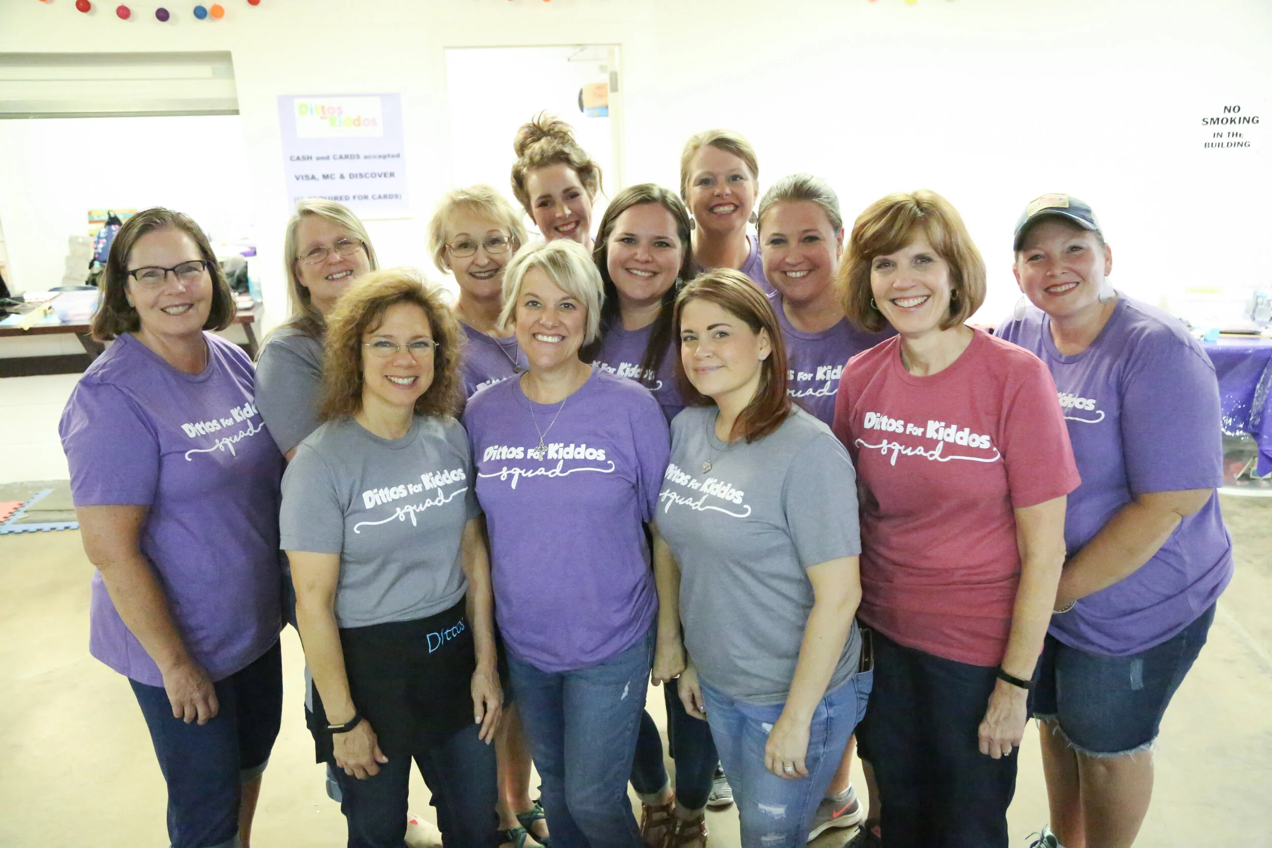 Group of women smiling, wearing purple and gray shirts with the text 'Ditos for Kiddos' on them, indoors at an event.