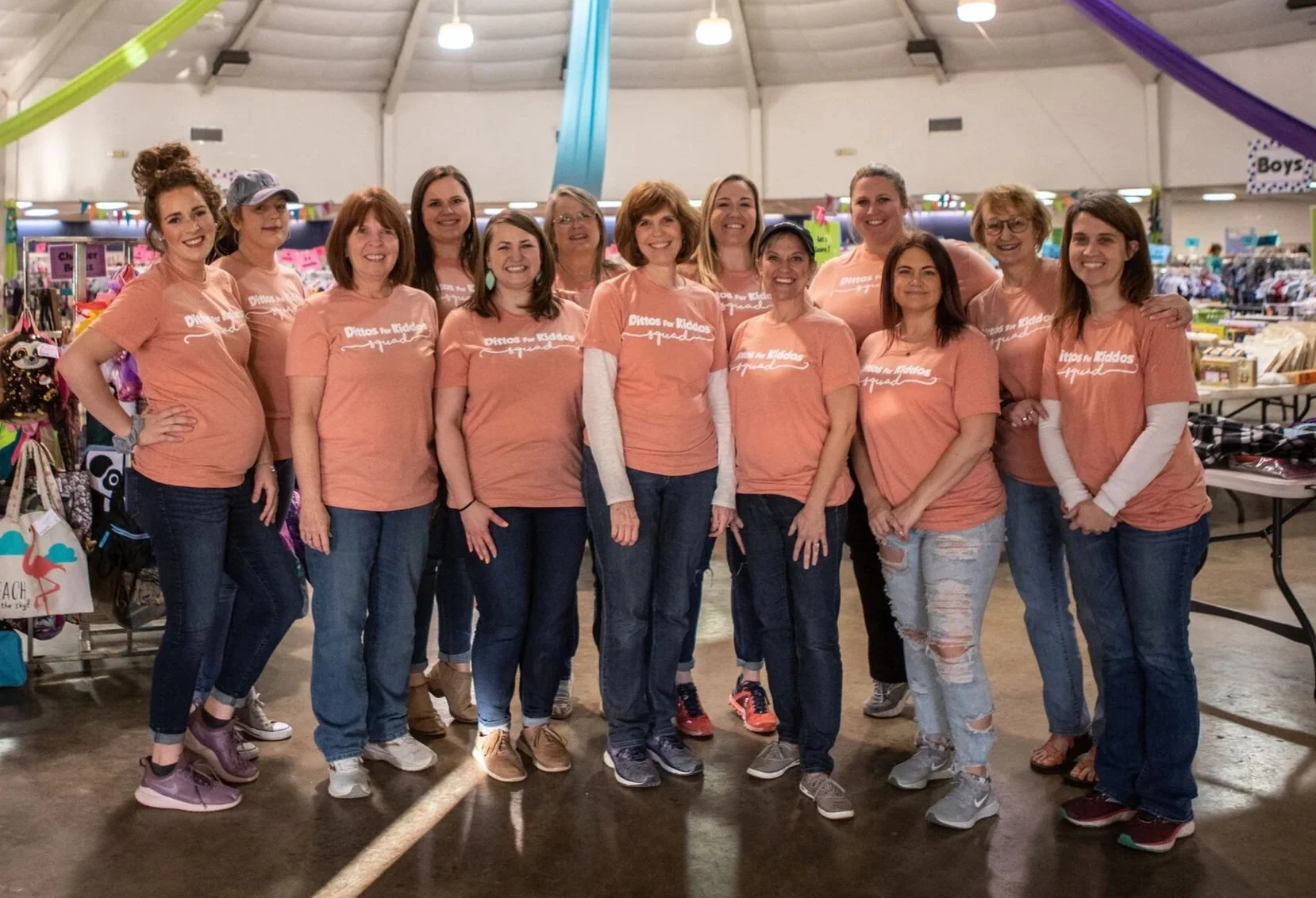 Group of women wearing matching peach T-shirts with the text 'Ditts for Kiddos' printed on them, standing together inside a large, well-lit indoor space, possibly a community center or event hall, with tables and booths in the background.