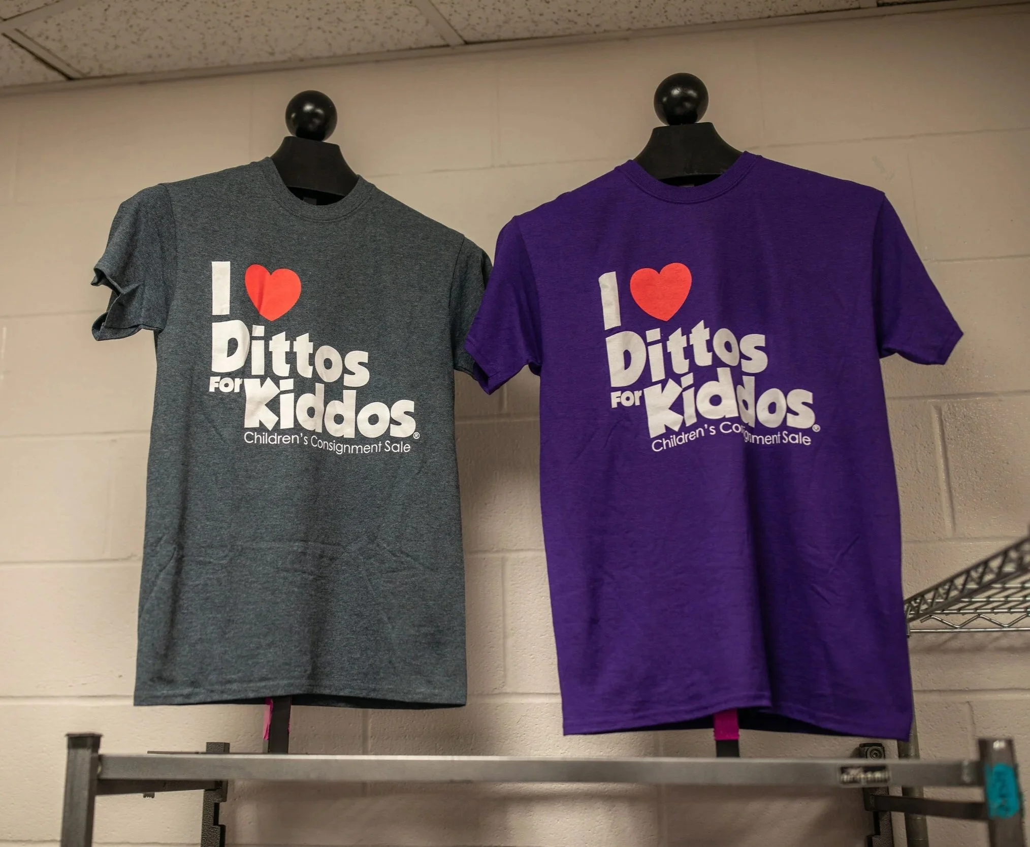 Two children's T-shirts hanging on black mannequin busts on a metal shelf. The T-shirts say 'I love PITTOS for Kiddos' and are gray and purple. It appears to be at a sale or donation event.