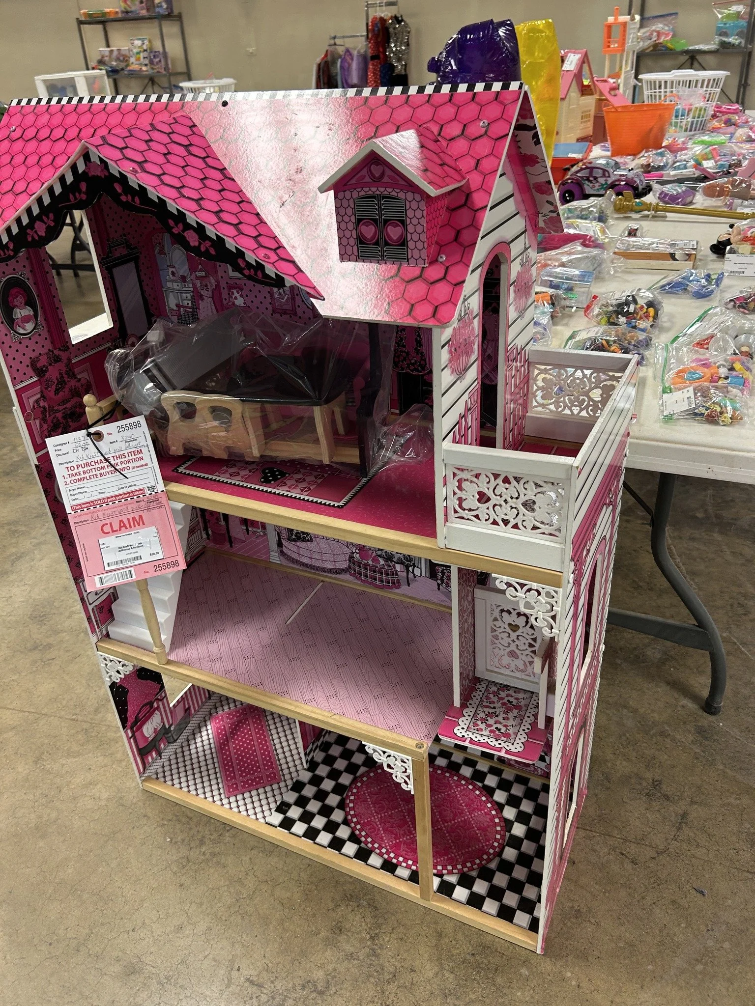 A pink and white miniature dollhouse with furniture and accessories inside, displayed on a table at a thrift store or sale event.
