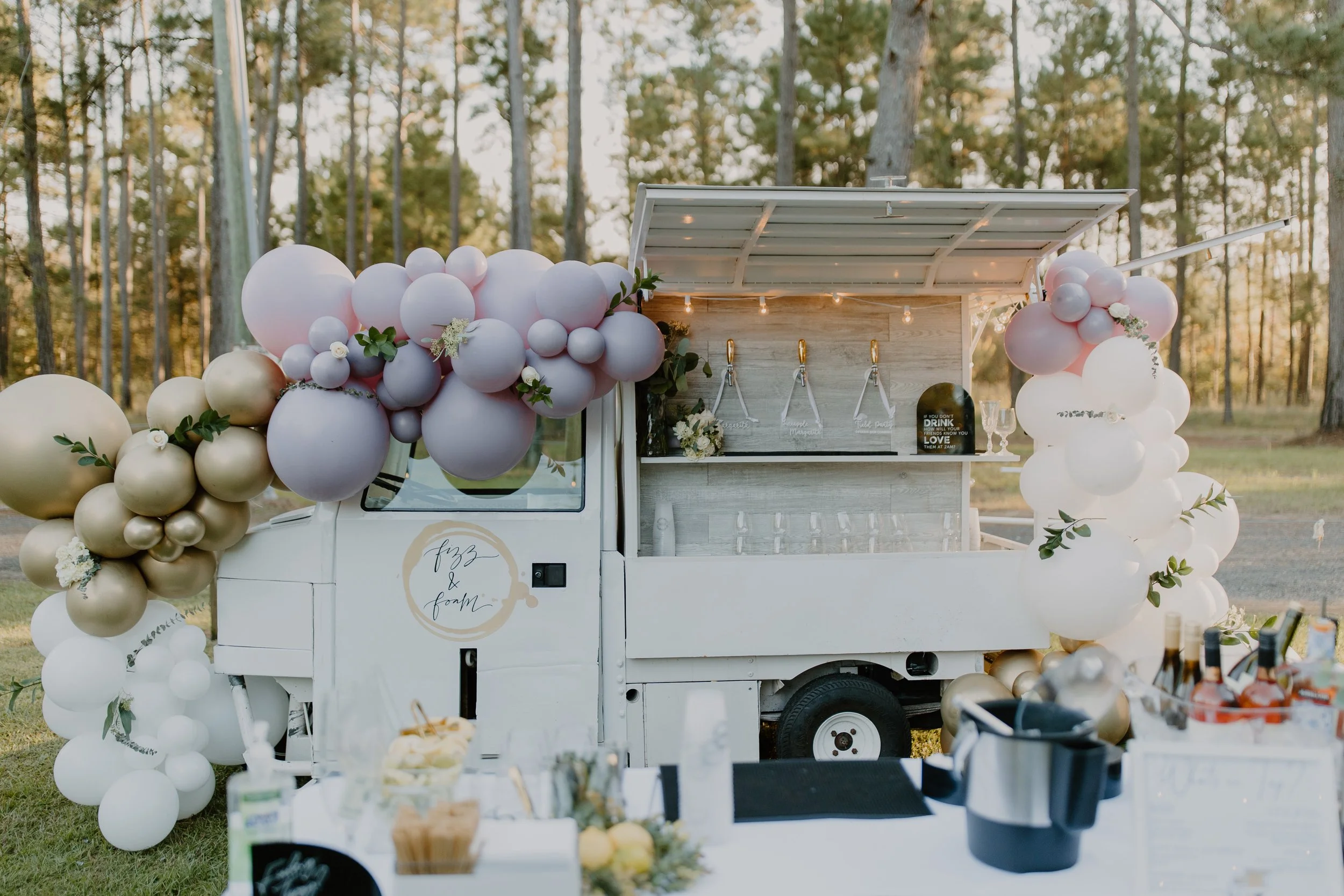 A white mobile bar decorated with pink, white, and gold balloons, set up outdoors in a wooded area for an event or celebration.