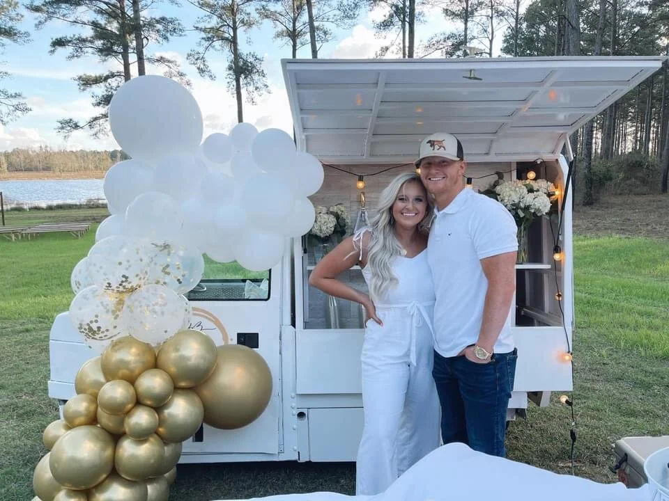 A smiling couple dressed in white standing in front of a small white food truck decorated with gold and transparent balloons, flowers, and string lights, outdoors near a lake and trees.
