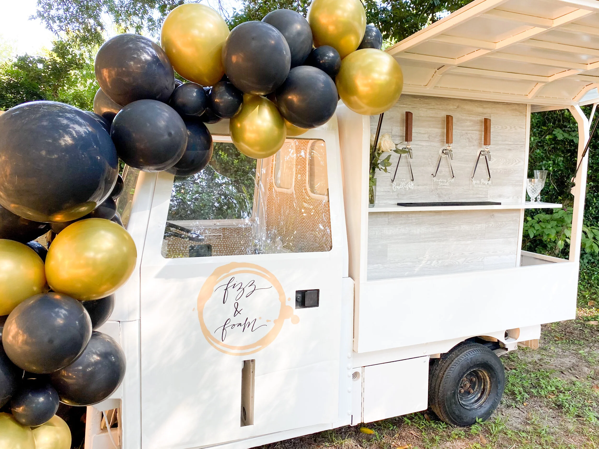 A small white mobile bar cart decorated with black and gold balloons, with a sign reading 'fizz & foam'. The cart has a built-in soda fountain and shelves holding a flower vase and glassware, outdoors on grass with trees in the background.