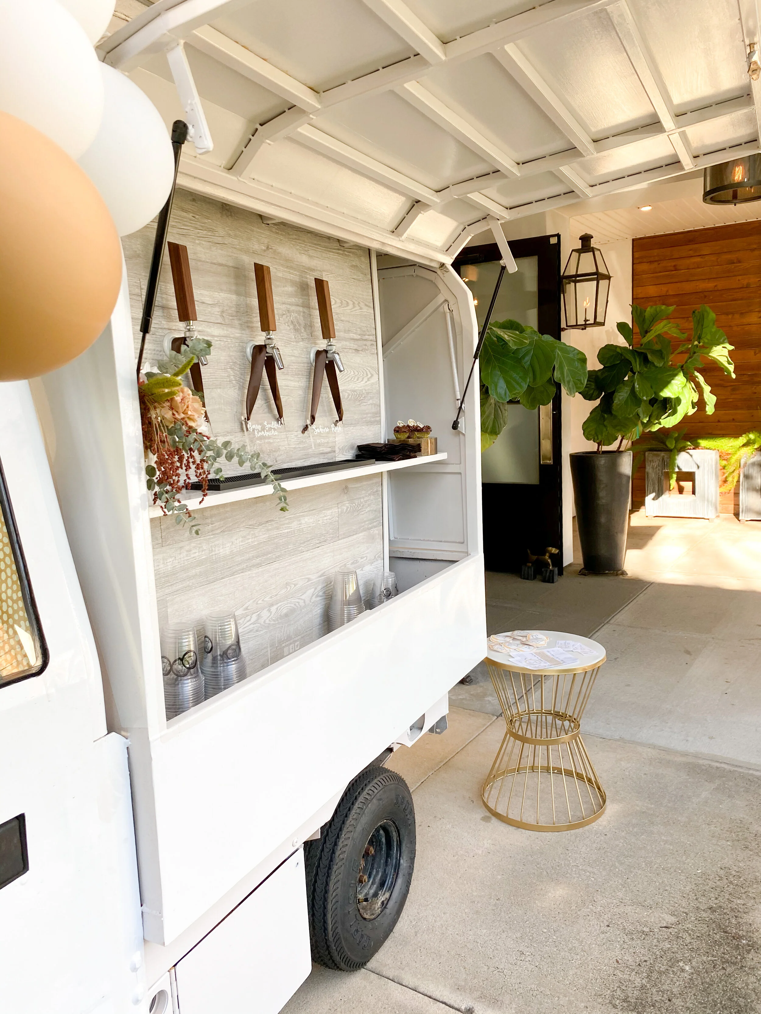 A white food truck with a serving window, blurred balloons on the left, and a small round gold wire table outside. In the background, large green plants and a lantern-style light fixture are visible.