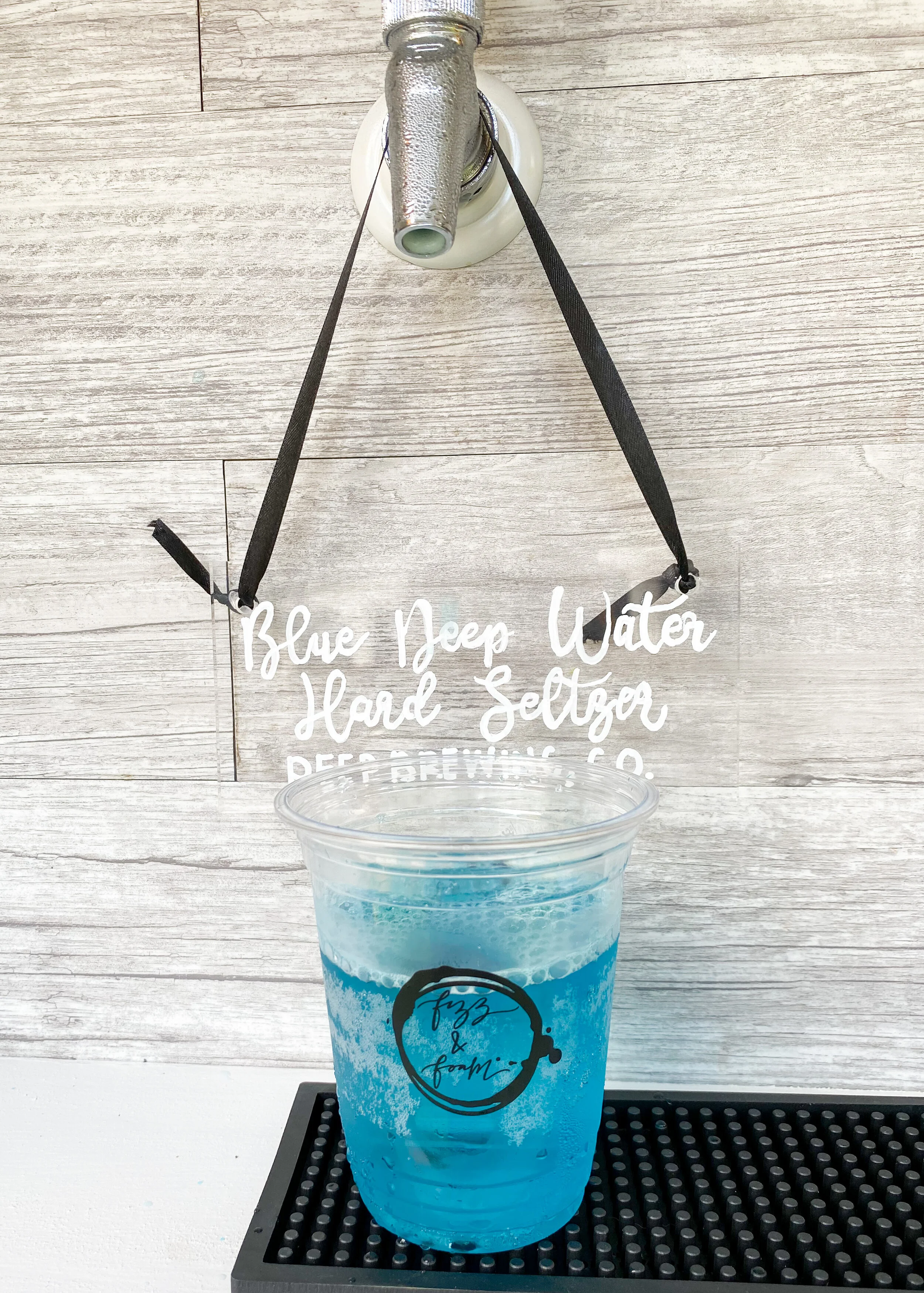 A clear plastic cup filled with blue liquid and foam on top, placed on a black rubber mat. In the background, there is a wooden wall with a sign hanging by black straps that reads "Blue Dee Water Hard Seltzer."