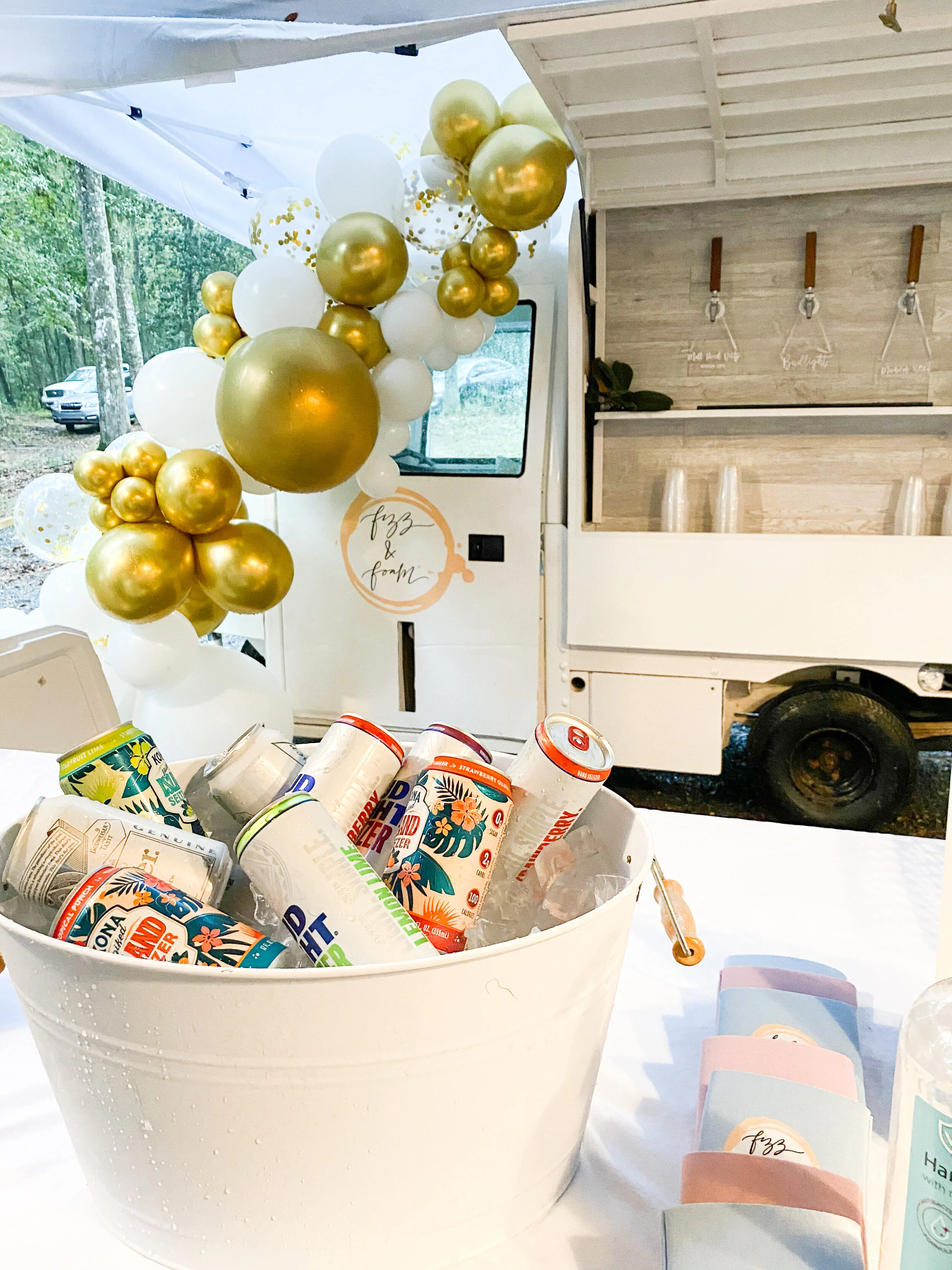 Decorated mobile bar with white, gold, and confetti balloons, cans of sparkling water and soda in a white bucket, with pink and blue napkins and a hand sanitizer bottle nearby.