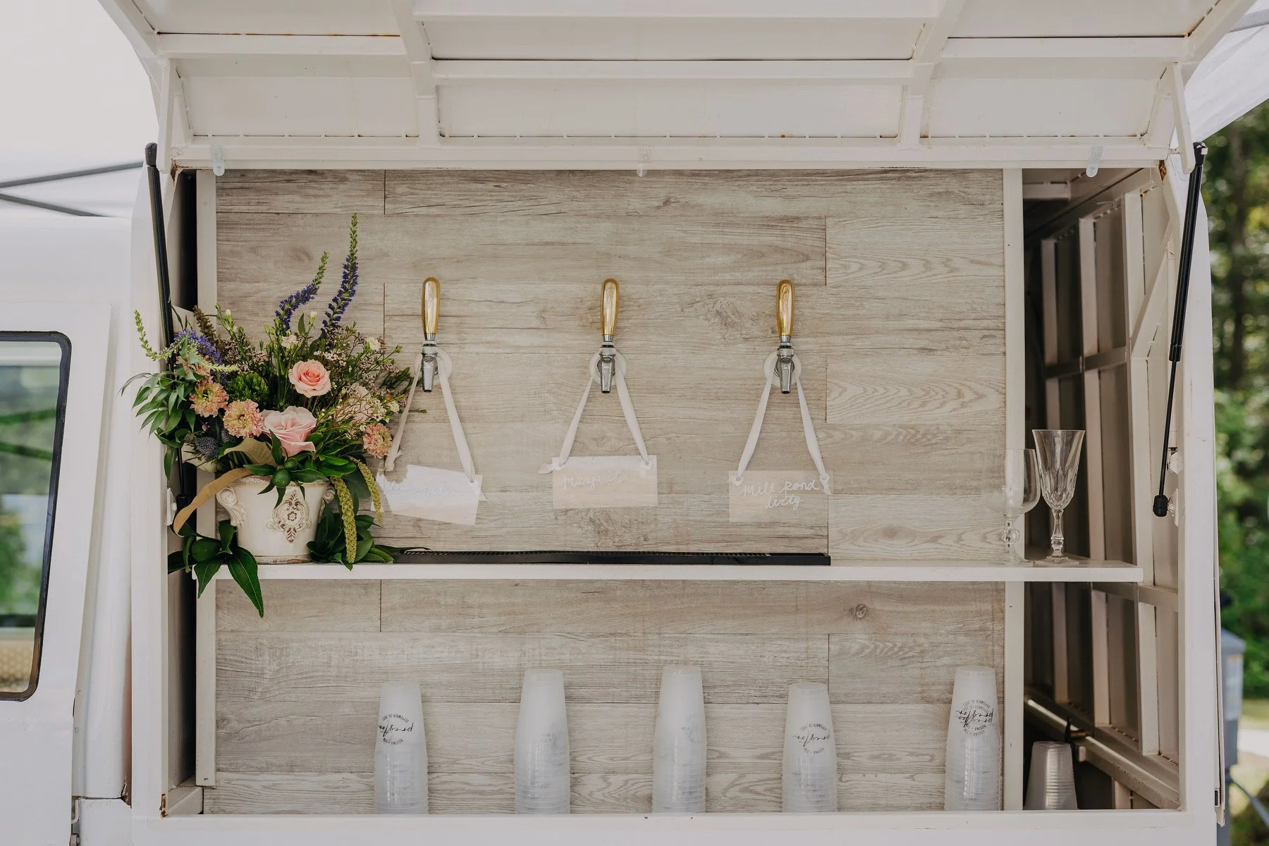 A mobile bar cart with three taps, a bouquet of flowers on the left, clear cups on the bottom shelf, and glasses on the right side, set outdoors.