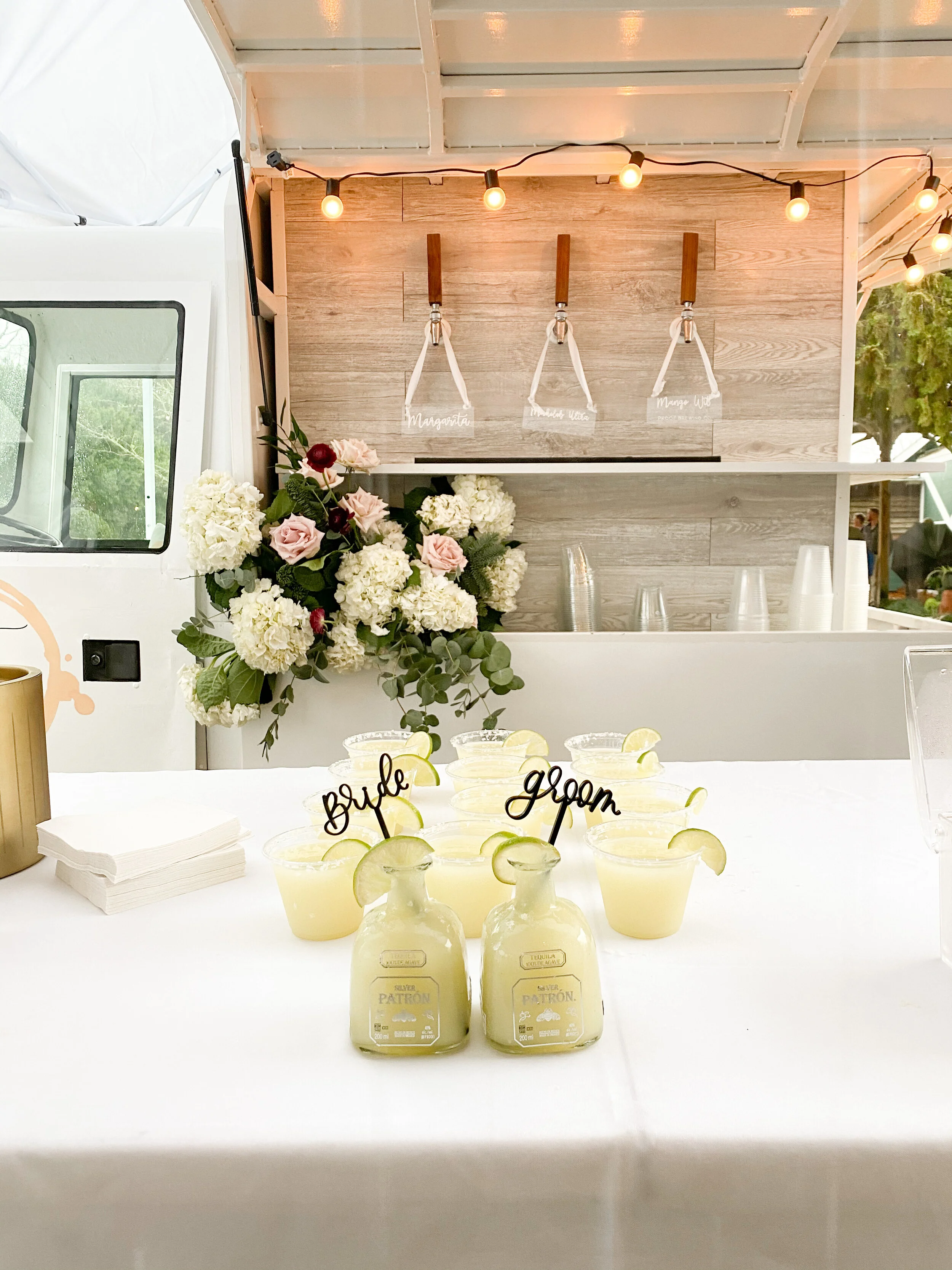 Table with six margarita drinks garnished with lime slices, two with 'groom' and 'bride' signs, floral arrangement of white and pink flowers behind, and a wooden bar area with hanging ingredient signs and string lights in the background.