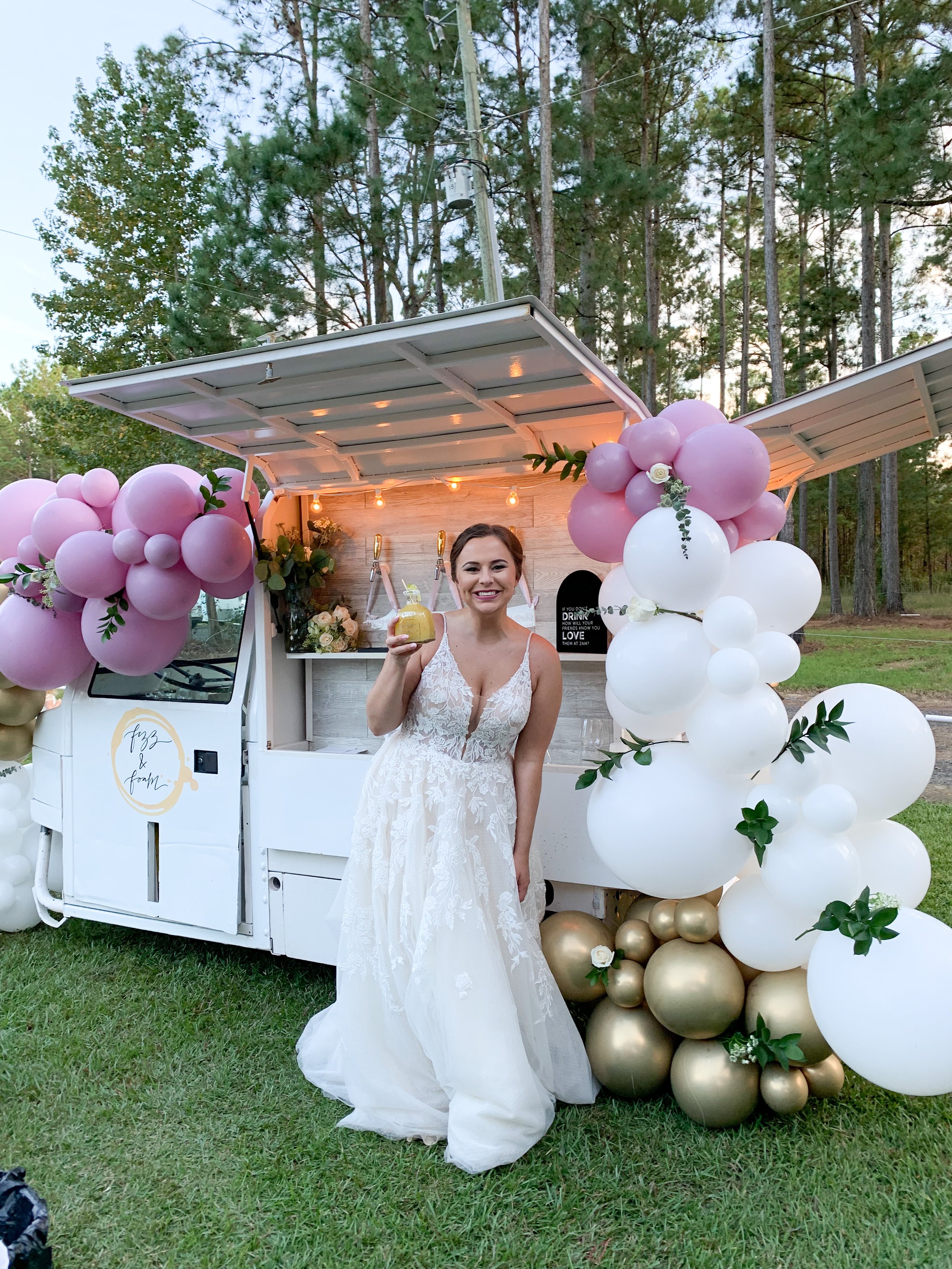 A smiling woman in a white wedding dress standing next to a decorated mobile bar with pink, white, and gold balloons at an outdoor event with trees in the background.